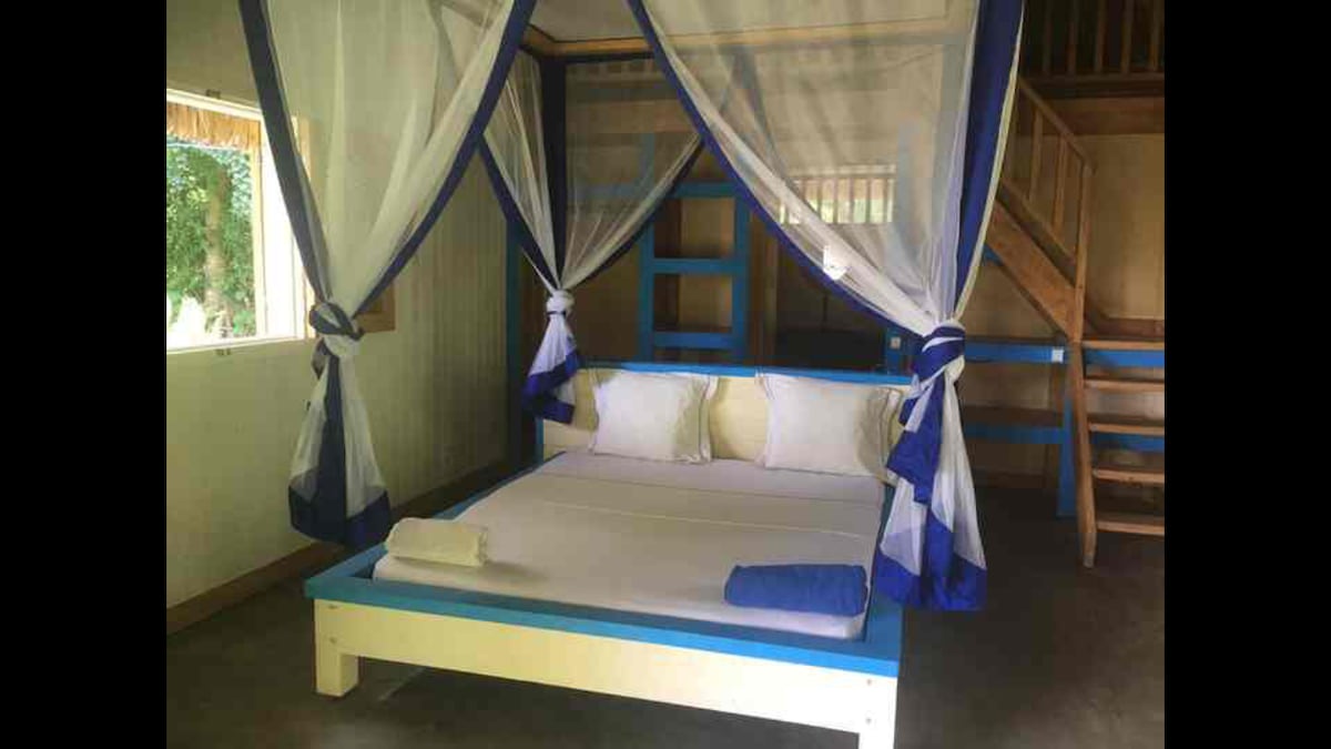 A sleeping area features a spacious bed with white linens, framed by canopy drapes in blue and white. Two pillows rest atop the bed, and a folded towel is placed at the foot. Natural light enters through the nearby window, revealing a wooden staircase in the background.