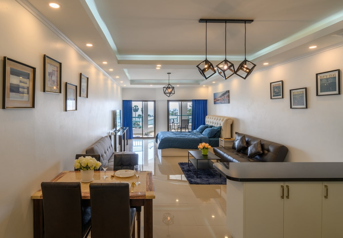 An open and airy living space features a cozy dining area with a table set for two, alongside a spacious kitchen counter. Two stylish sofas, one light and one dark, complement the room. Large glass doors provide access to a balcony with a view.