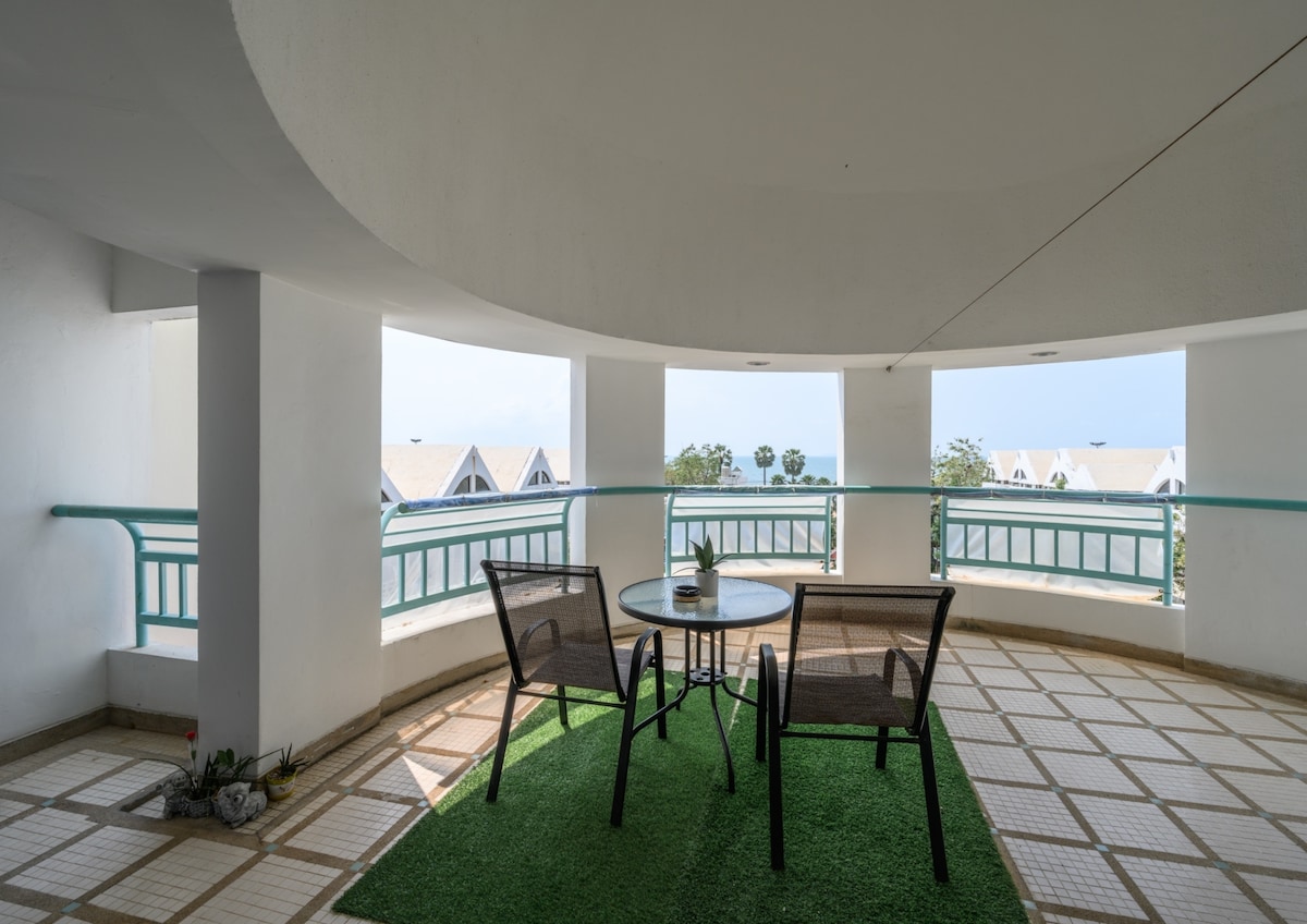 A spacious balcony area features a circular design, surrounded by large windows offering views of the beach. A round table and two chairs are set on a grassy mat, inviting relaxation while enjoying the fresh sea breeze.