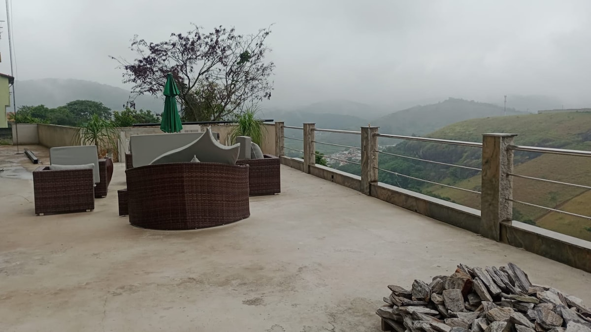 An outdoor terrace is visible, featuring woven seating arrangements that invite relaxation. A green umbrella provides shade, while a stack of wood is neatly arranged nearby. The backdrop showcases misty hills, creating a serene and tranquil environment.