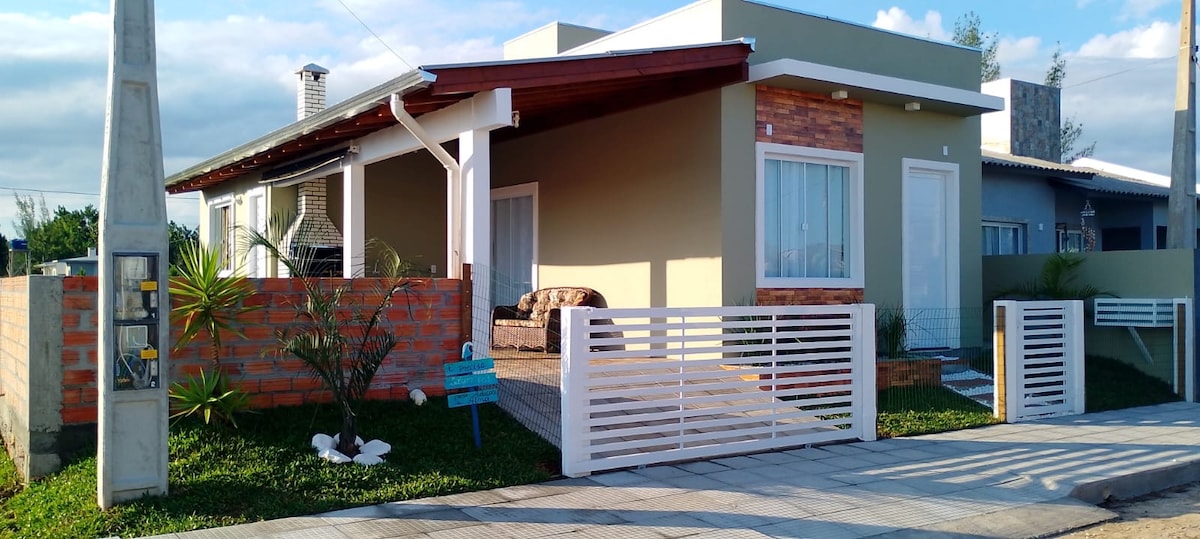 A modern exterior view of the house is shown, featuring a welcoming porch with comfortable seating. The facade includes a combination of stone and light-colored walls. Lush greenery surrounds the entrance, and a decorative fence provides a clear boundary while allowing visibility of the inviting walkway.