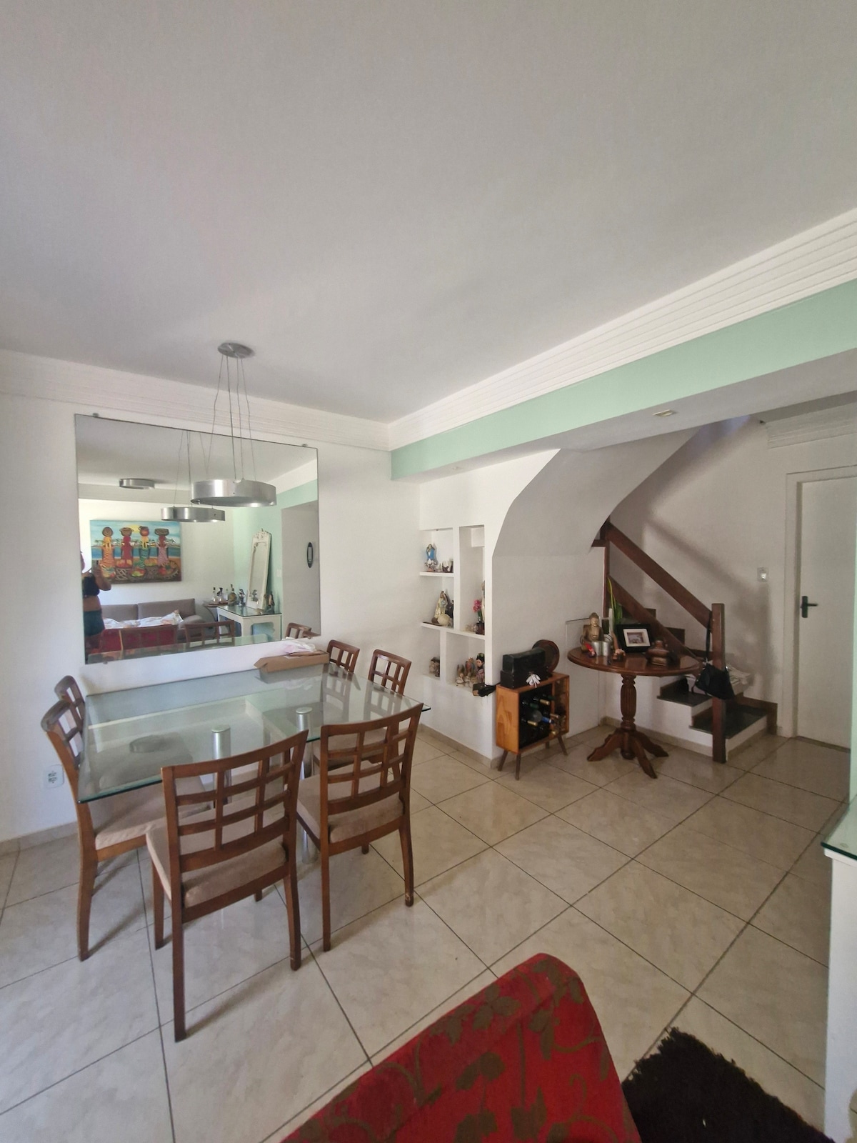 A dining area features a glass table surrounded by six wooden chairs, placed on a tiled floor. A staircase is visible in the background, leading to an upper level. The adjacent wall showcases built-in shelves, enhancing the space's functionality.