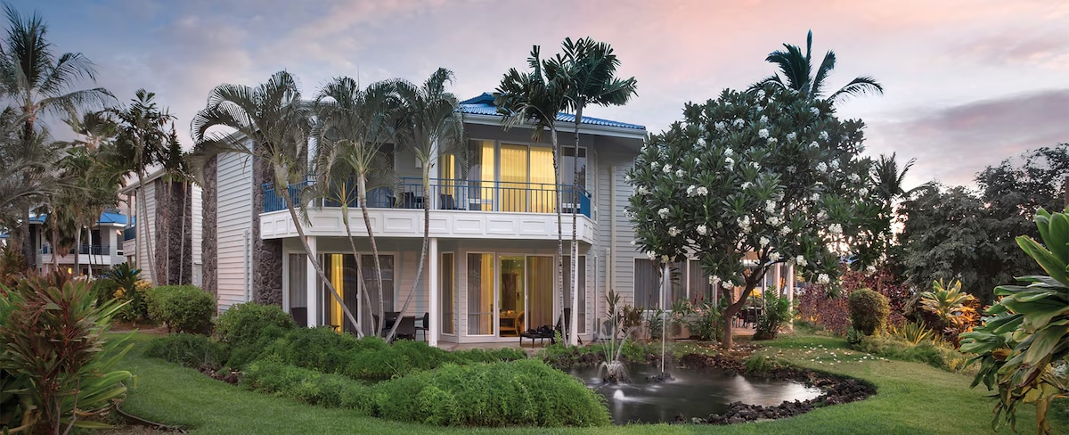 The exterior of a two-story building is showcased against a colorful sunset. Lush greenery surrounds the property, with various palm trees and flowering plants. A serene pond with a small water feature is visible in the foreground, enhancing the tranquil environment.