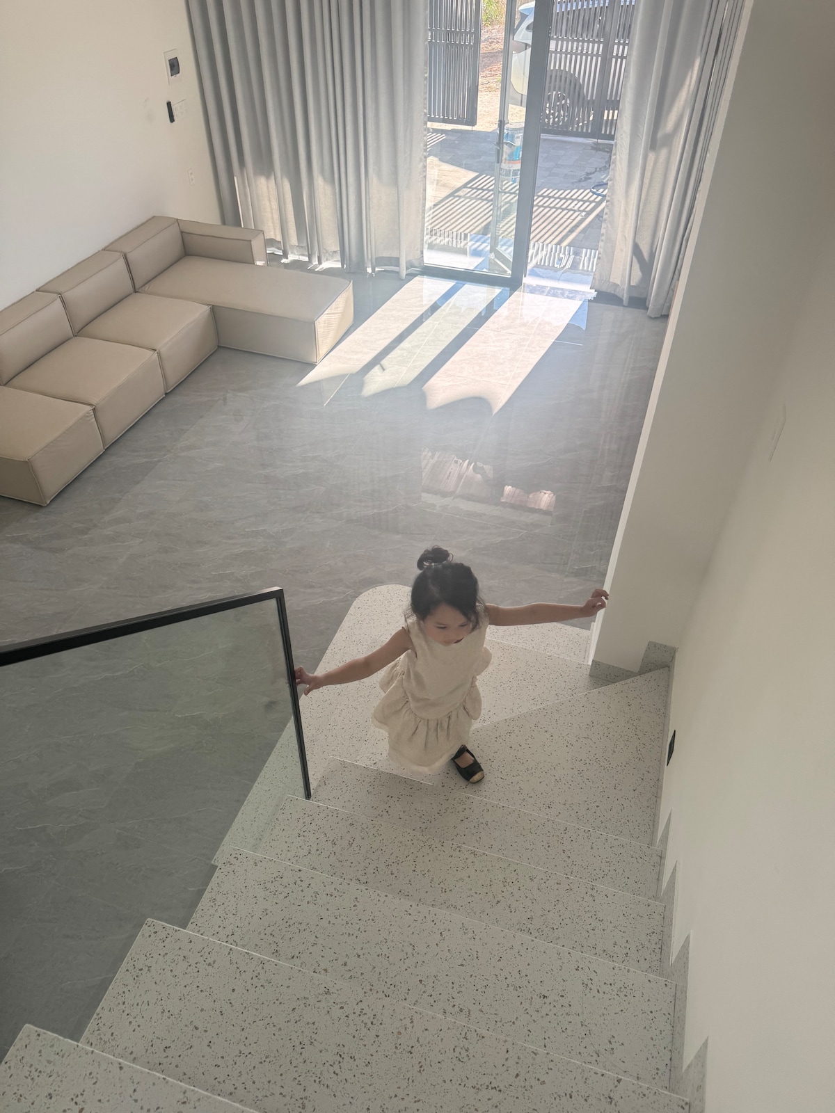 A child is seen descending a sleek staircase. The space is filled with natural light, highlighting the smooth, light-colored flooring. A modern, sectional couch is positioned in the background, accompanied by floor-to-ceiling curtains that partially frame windows opening to an outdoor area.