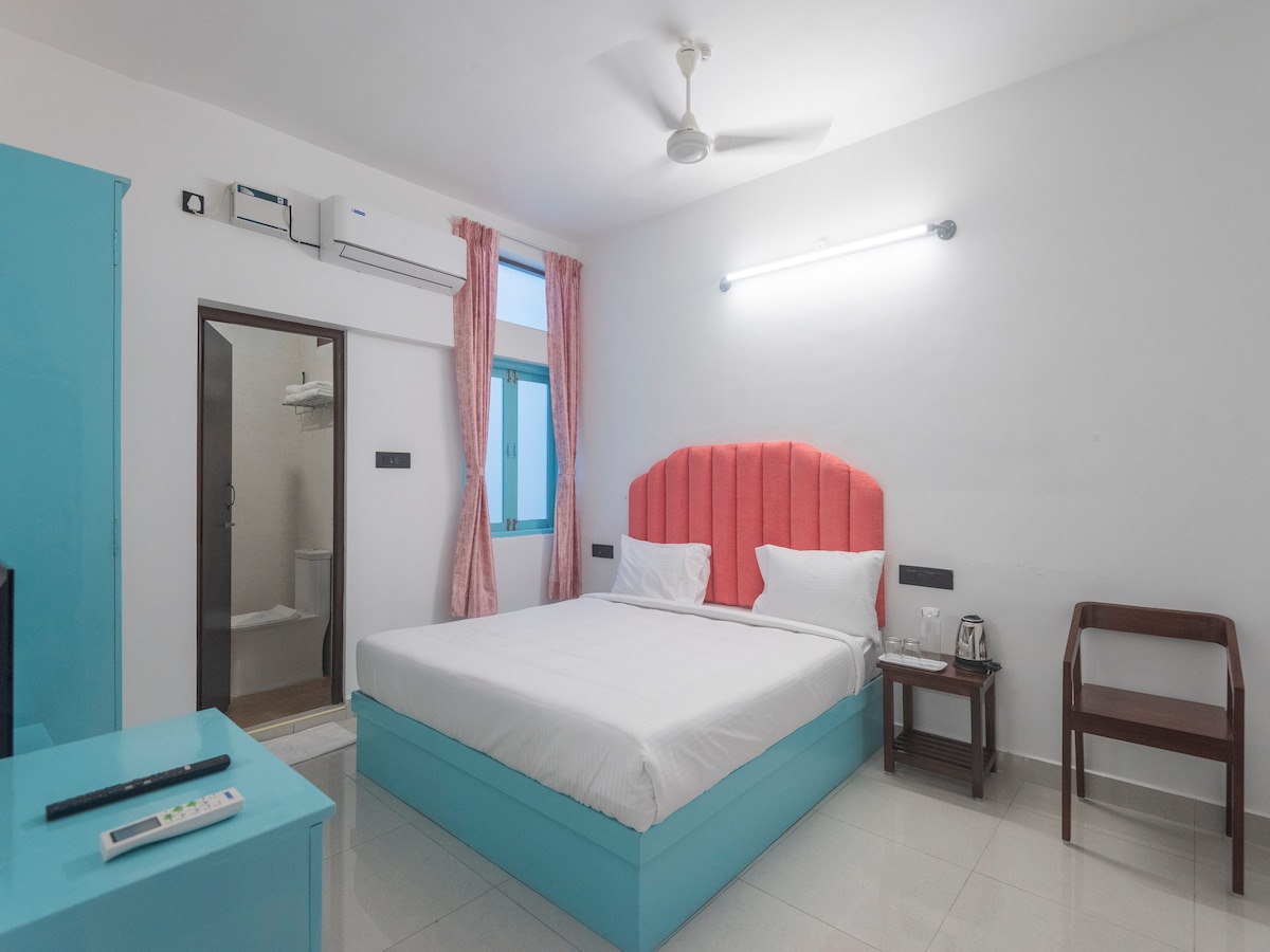 A bright guest room features a spacious bed with a coral headboard and crisp white linens. A blue wardrobe and bedside table complement the decor. Natural light enters through a window with light curtains, while an en-suite bathroom is visible through an open door.