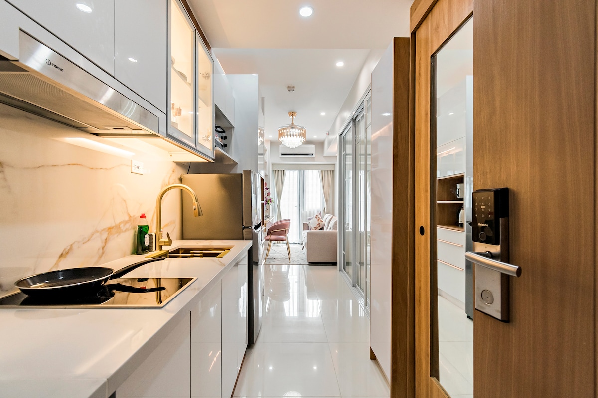 A modern kitchen with sleek cabinetry and a polished countertop is visible. Appliances include a stovetop and an oven, with a frying pan on the burner. The space is well-lit with natural light flowing from the adjacent area, leading to a living room with a chandelier.