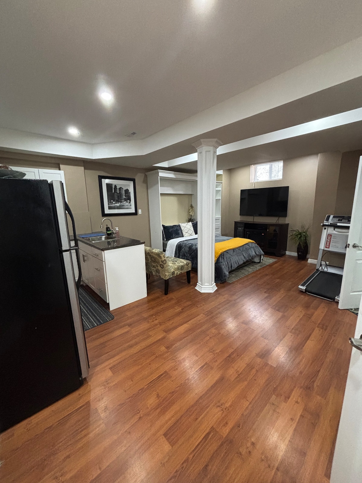 A spacious basement studio is observed, featuring warm wood flooring. A cozy queen size bed is positioned against a wall, complemented by a television across the room. A compact kitchen area offers a refrigerator and cabinets. A chair is placed nearby, with a treadmill visible in the corner.