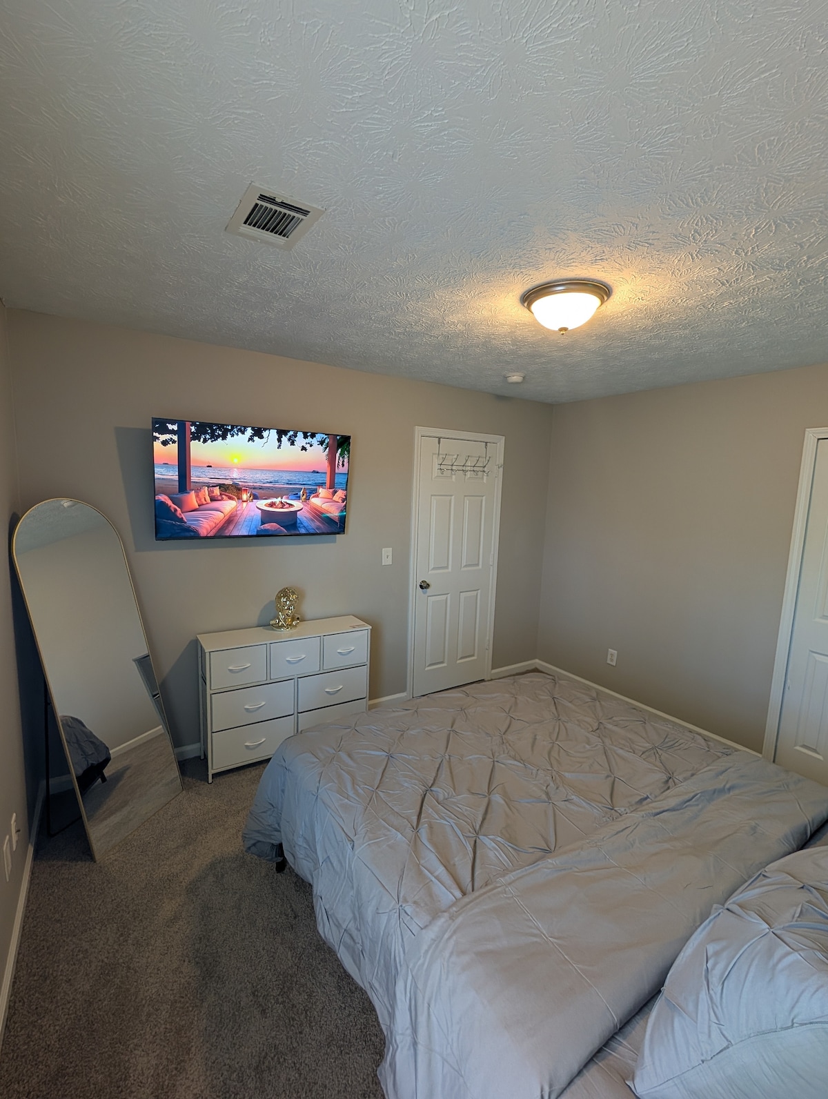 A cozy bedroom features a spacious bed covered in a light gray comforter. A flat-screen television is mounted on the wall, displaying a serene landscape. A tall mirror stands in the corner, and a white dresser is positioned against the wall, providing ample storage.
