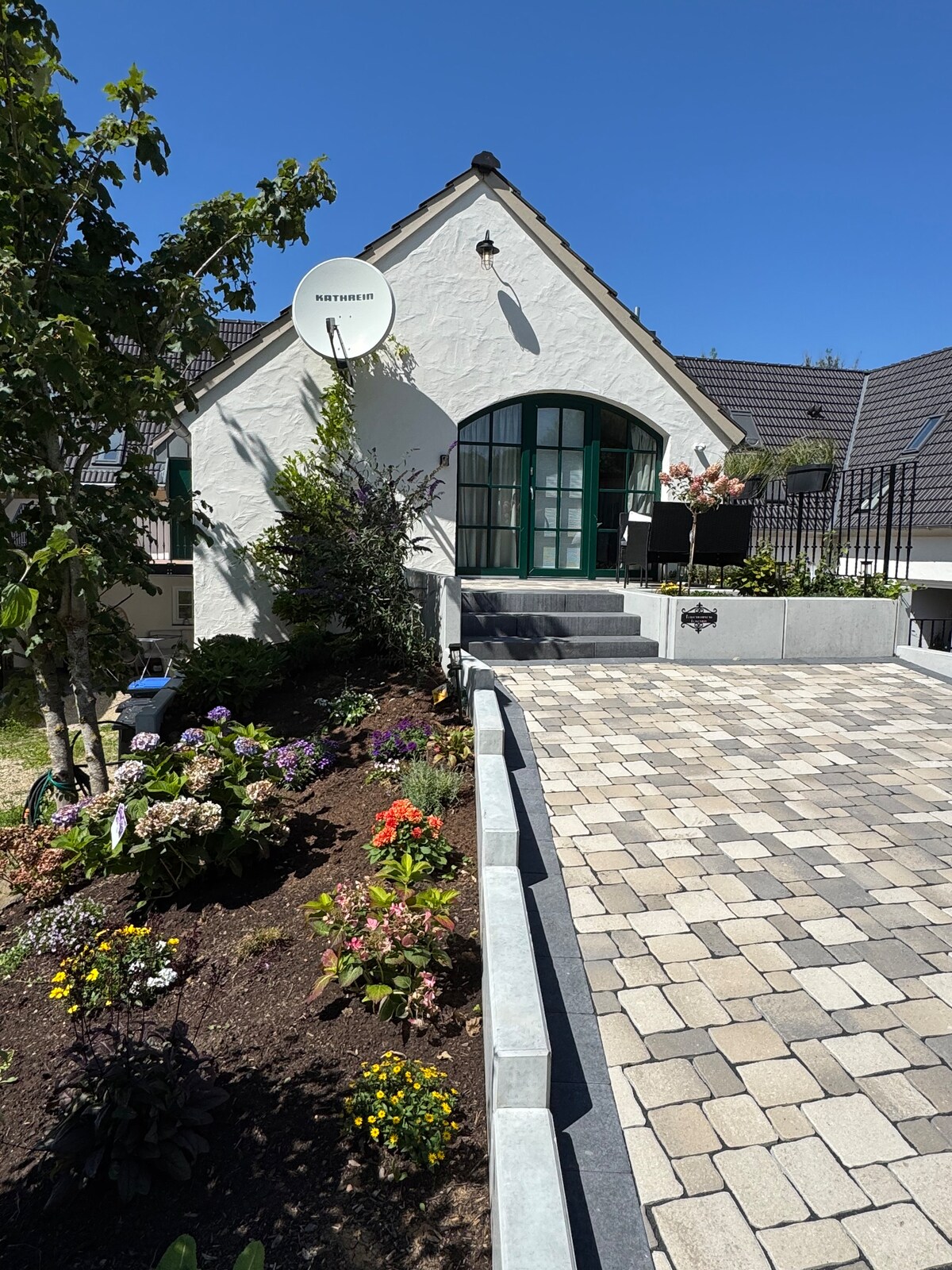 The exterior of a renovated cottage is presented, featuring a charming entrance accented by an arched door. Lush flower beds with vibrant blooms line the pathway, while a cobblestone driveway leads up to the residence. Leafy trees provide natural shade, enhancing the outdoor space.