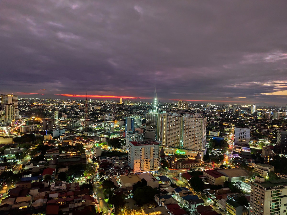 A panoramic view of the city skyline is presented at twilight, with a mix of residential and commercial buildings illuminated by city lights. The horizon showcases a blend of colors as the sun sets, offering a glimpse of the vibrant urban atmosphere.