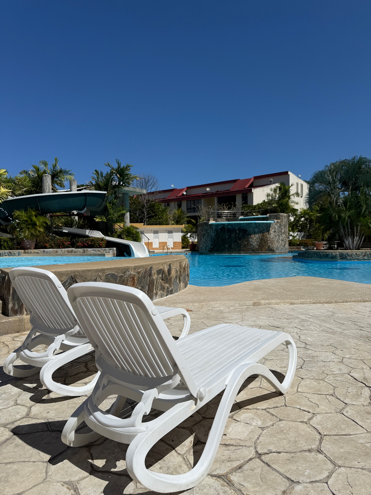 Two white lounge chairs are positioned beside a large swimming pool. The pool's clear water reflects the blue sky, while a waterfall feature adds to the serene ambiance. Lush greenery and a multi-level building are visible in the background, enhancing the relaxing atmosphere.