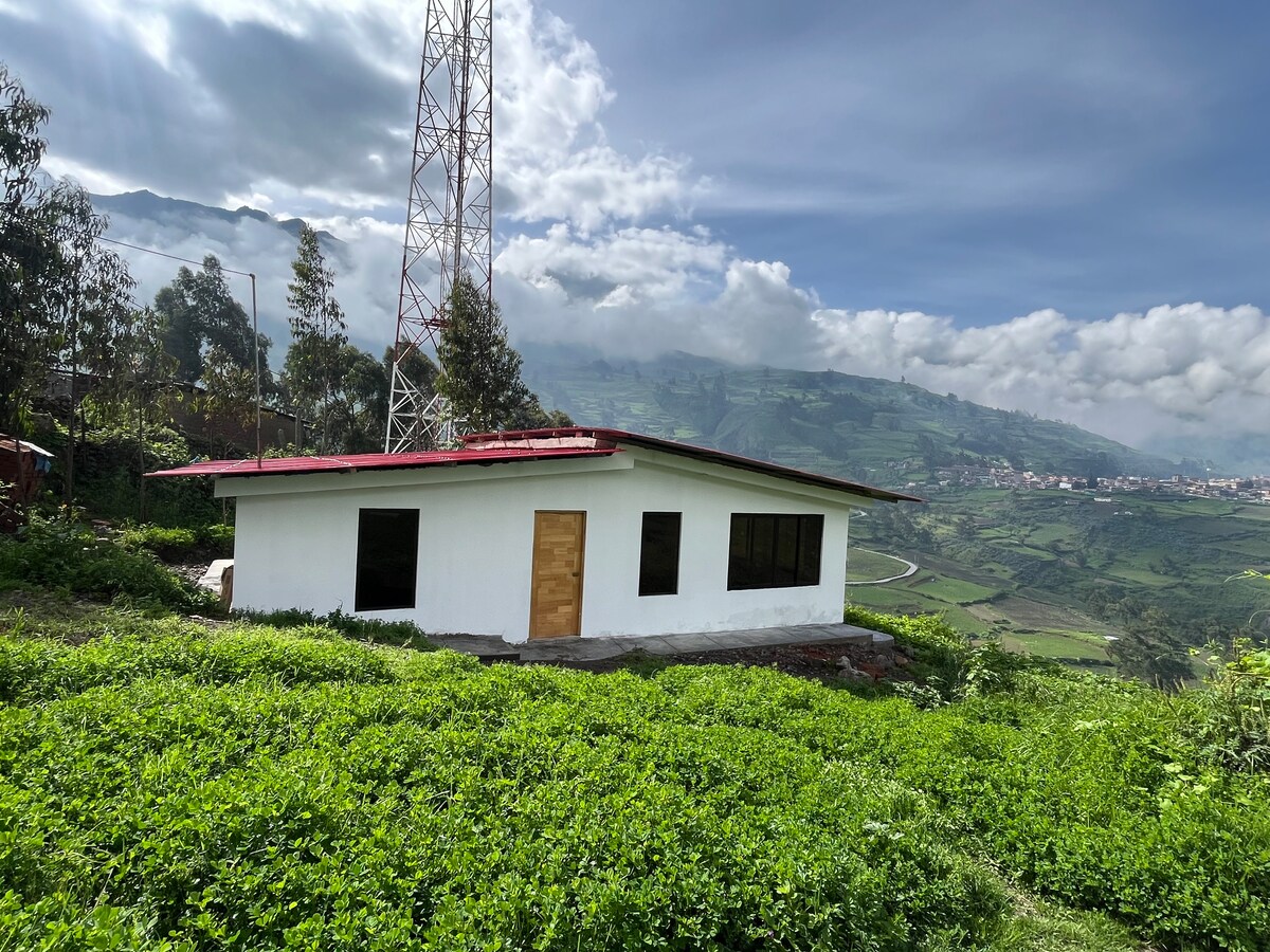 A simple, single-story building is surrounded by lush green foliage, with expansive windows providing views of the rolling hills and distant mountains. A communication tower stands nearby. The sky is partly cloudy, contributing to a serene landscape.