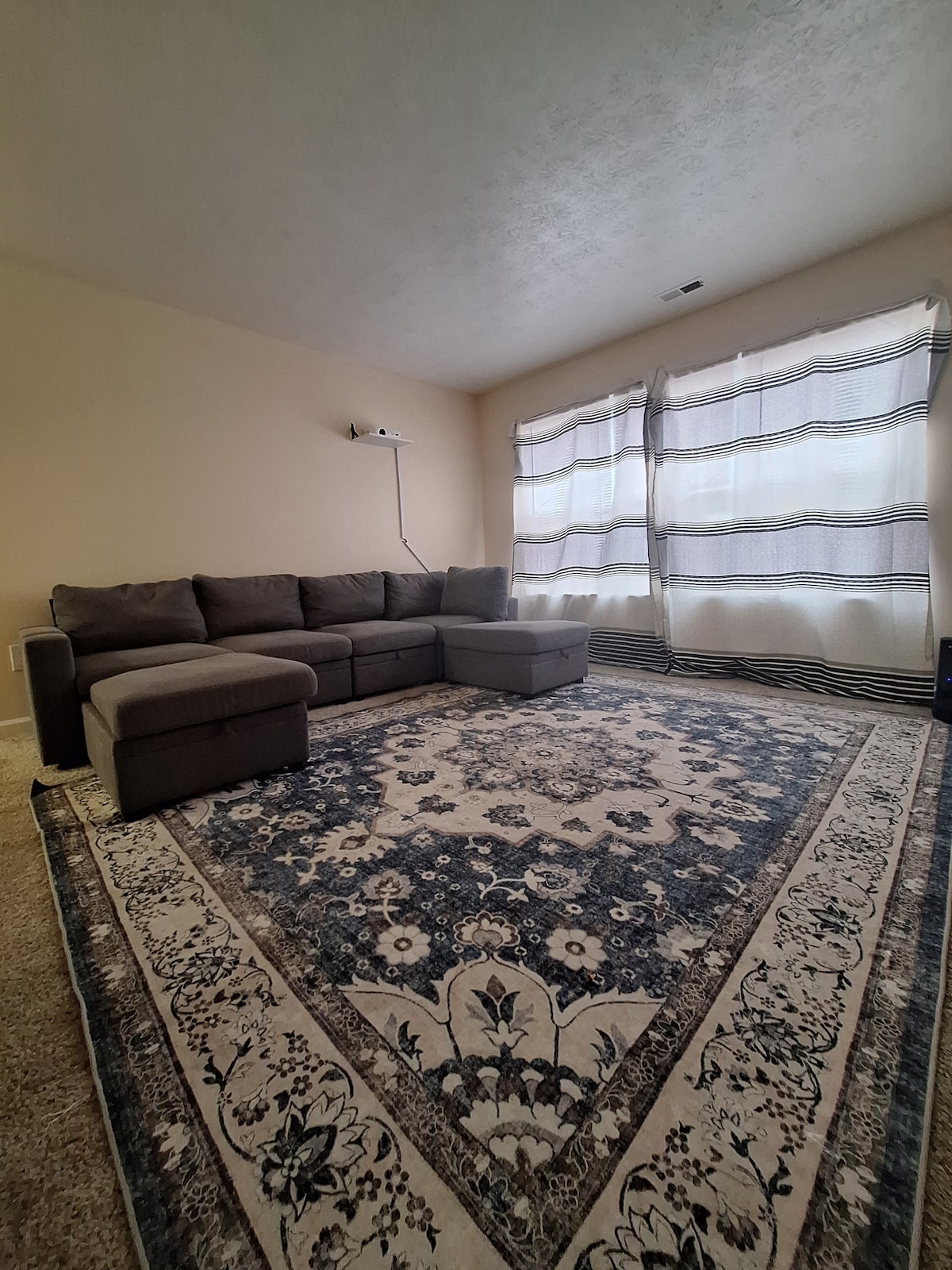 A cozy living area features a large sectional sofa paired with a matching ottoman. Natural light filters through sheer curtains covering the wide windows. A decorative area rug with intricate designs enhances the space, providing a comfortable setting for relaxation.