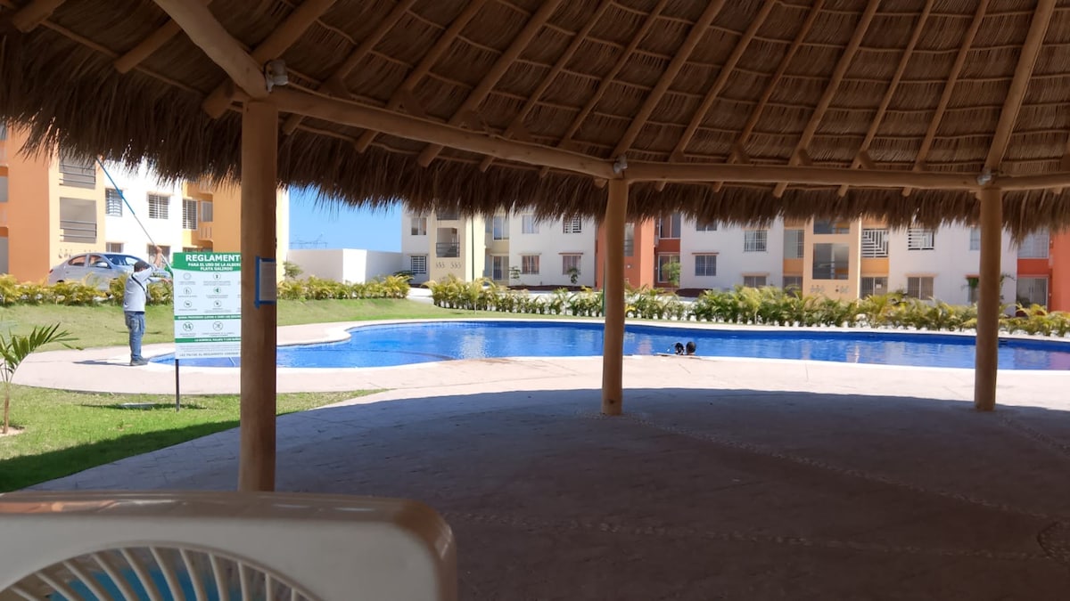 A shaded seating area is seen under a thatched roof, offering a view of a large swimming pool surrounded by landscaped greenery. Soft sunlight reflects off the water while colorful residential buildings provide a welcoming backdrop.