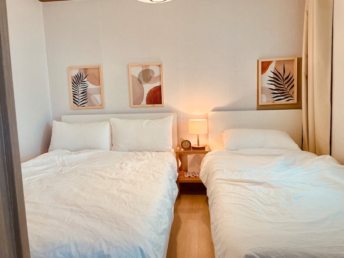 A welcoming bedroom features two spacious beds dressed in white bedding. Soft lighting illuminates the room from a bedside lamp on a small table. Abstract art pieces decorate the walls, adding subtle color and visual interest. A warm, neutral palette enhances the inviting atmosphere.