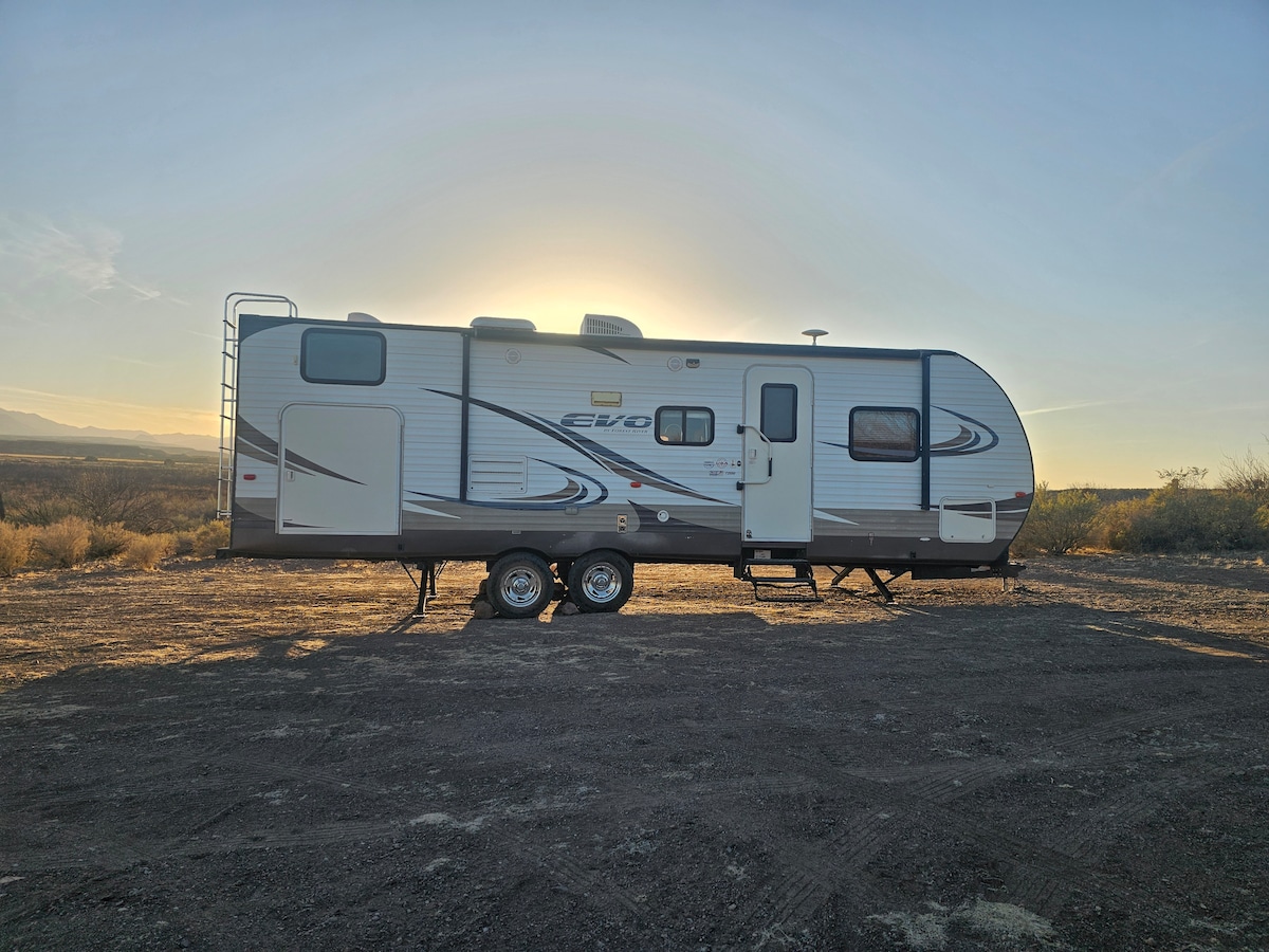 A travel trailer is positioned on a gravel area, with the sun rising behind it. The exterior features a streamlined design with a combination of neutral tones and subtle graphics, highlighting the entry steps and window placements.