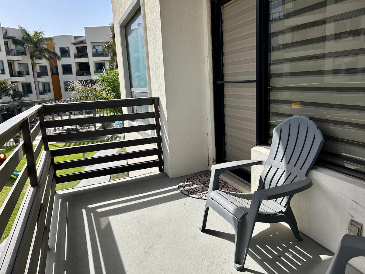A spacious balcony is presented with a light concrete floor and a single gray adirondack chair. Sunlight casts shadows through the balcony's slatted railing, overlooking green grass and well-maintained landscaping visible in the distance.