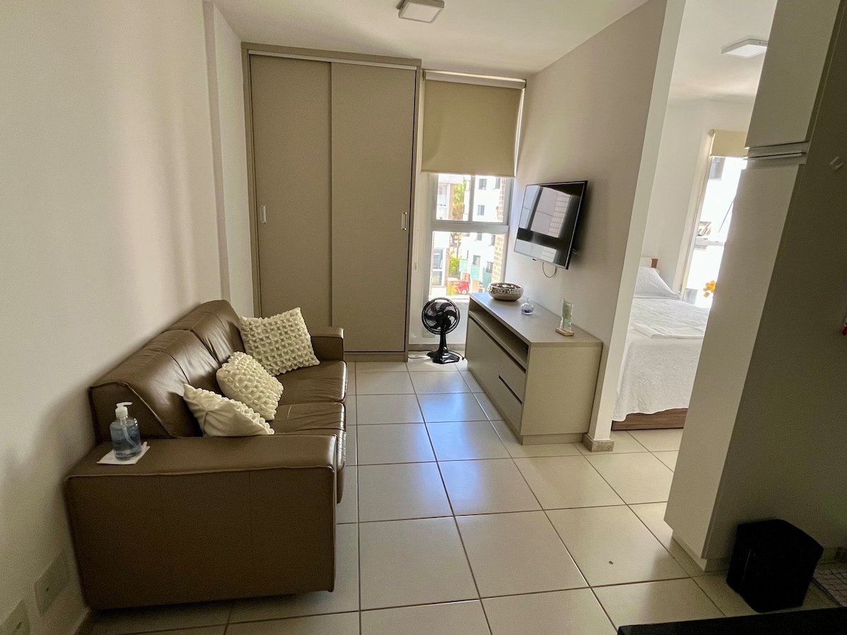A living area features a comfortable brown sofa adorned with decorative pillows. A wall-mounted television is positioned above a minimalistic cabinet, while a fan is placed on the floor. Natural light enters through a large window, and sliding doors provide access to additional storage.