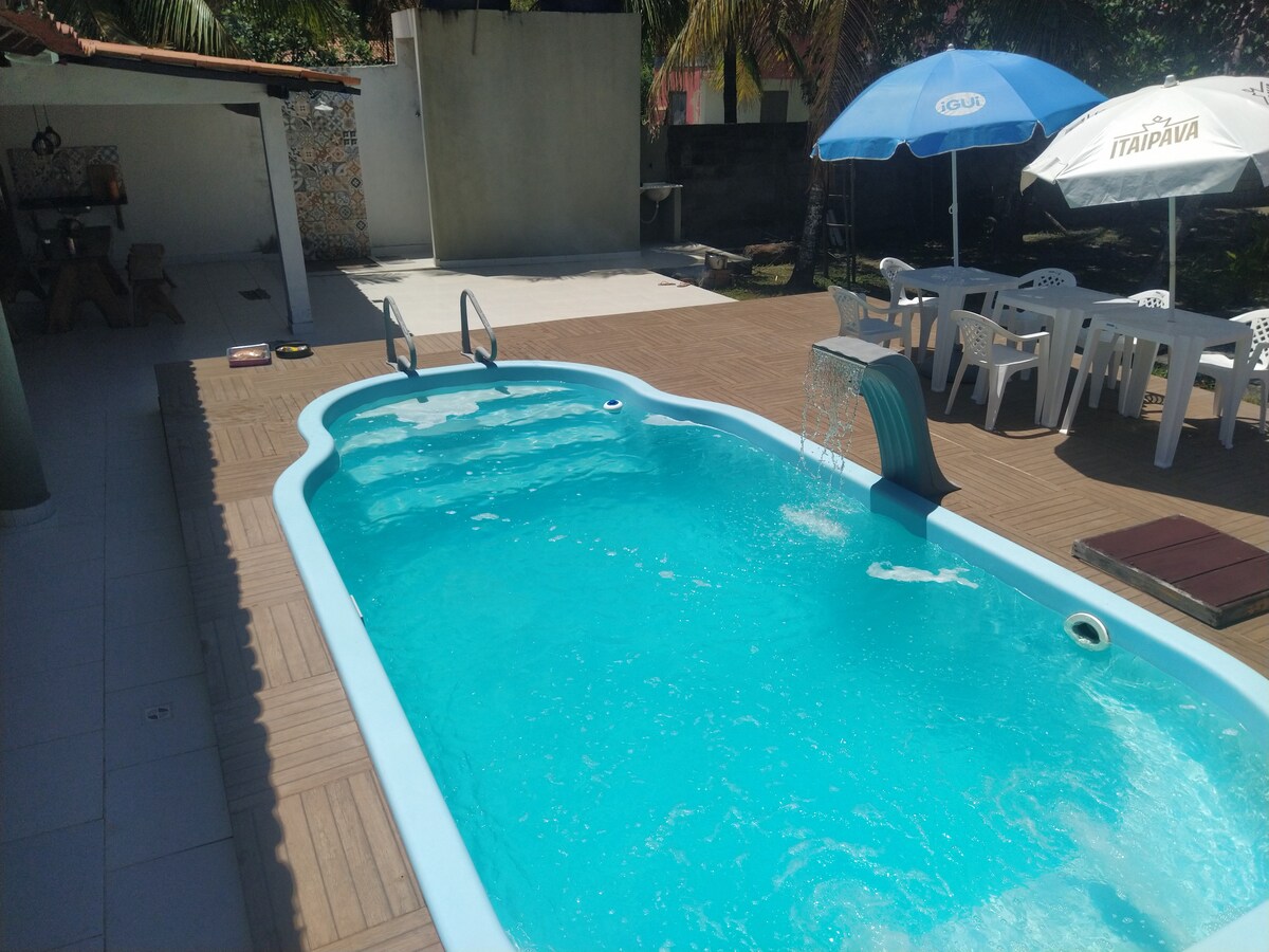 A refreshing swimming pool glistens under sunlight, featuring a water feature that creates a soothing sound. Surrounding the pool are sun loungers and a shaded area provided by umbrellas, offering comfort in the outdoor space. A spacious deck surrounds the pool, enhancing the inviting atmosphere.