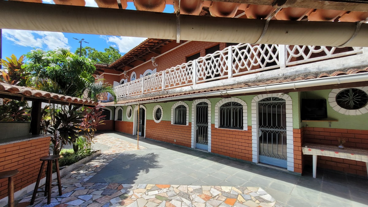 A quiet outdoor area is surrounded by lush greenery and features a mosaic stone pathway. The building showcases a two-story design with multiple arched windows, elegant railings, and a covered terrace. Sunlight filters through the trees, creating a bright and welcoming environment.
