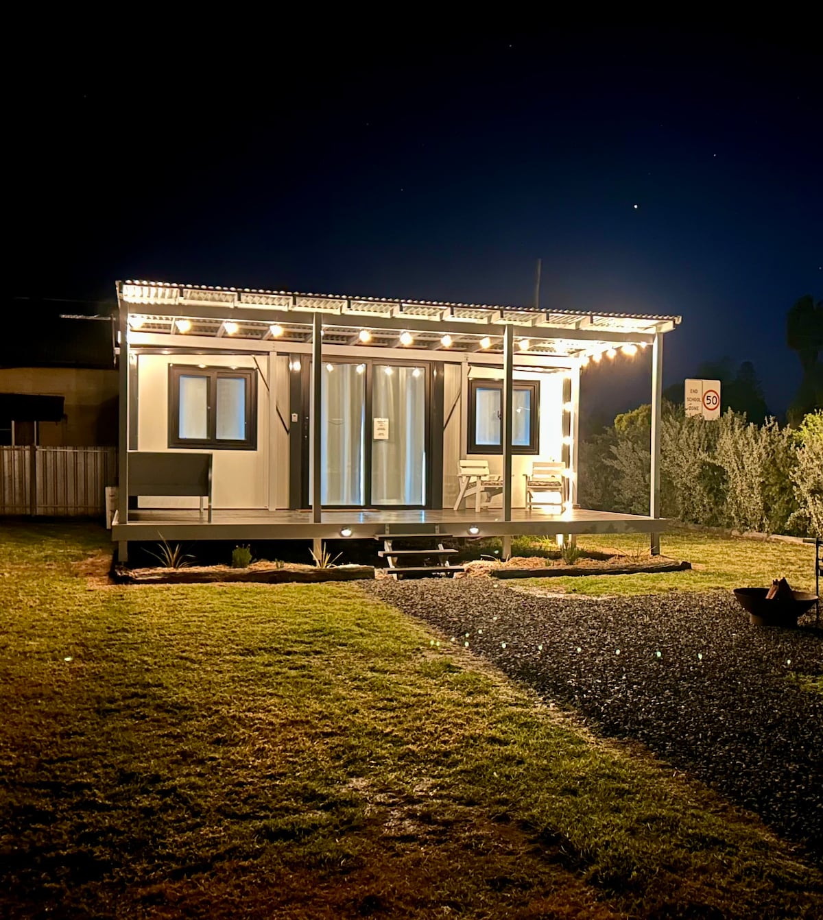 A cozy house is illuminated by soft string lights under a night sky. The front porch features two chairs, creating an inviting space. A gravel pathway leads to the entrance, surrounded by a lawn and shrubs, enhancing the outdoor atmosphere.