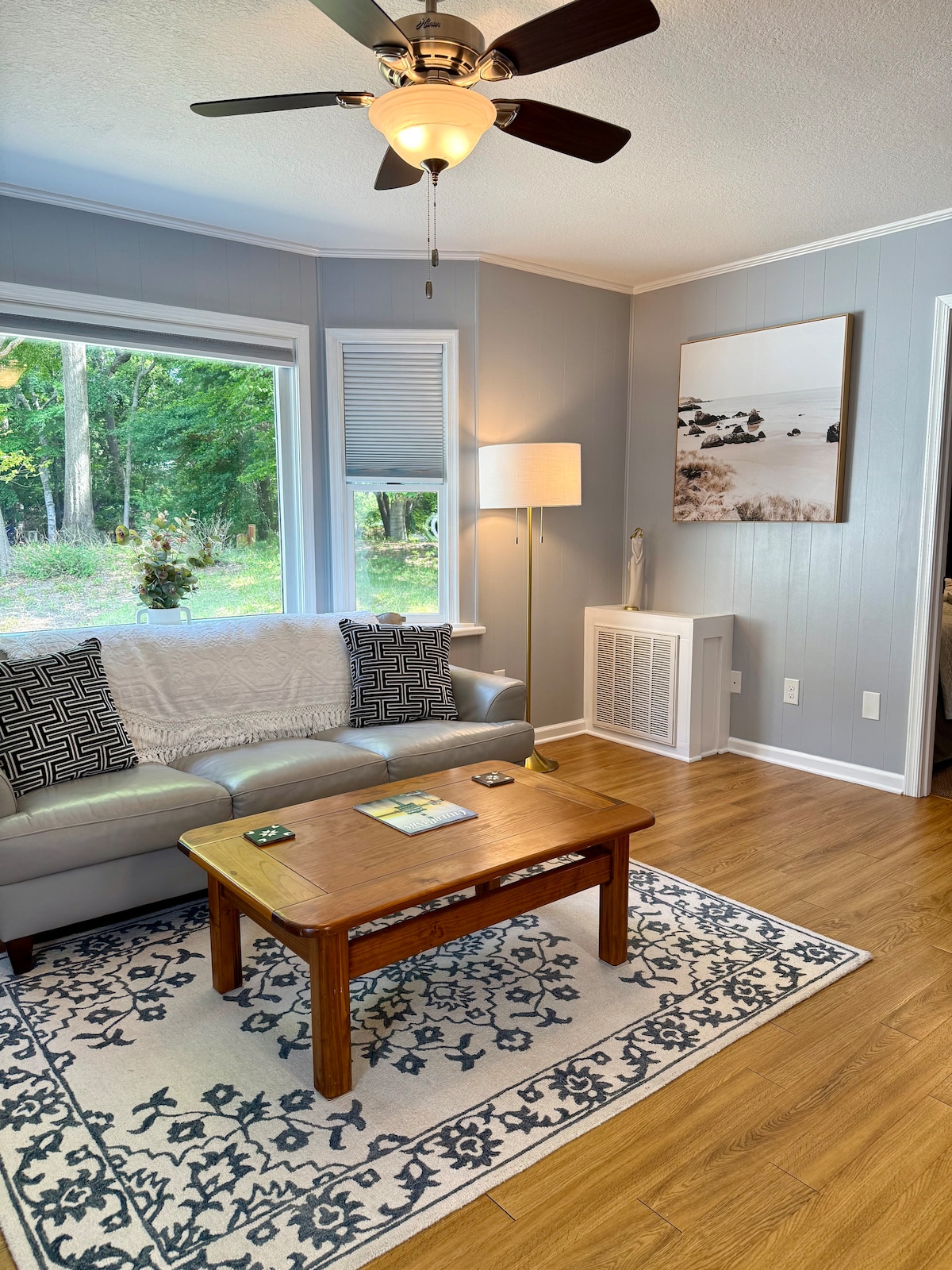 A comfortable living area features a light-colored sofa adorned with a textured throw. A wooden coffee table sits in front, complemented by a patterned area rug. A ceiling fan provides airflow, while large windows offer views of the surrounding nature.
