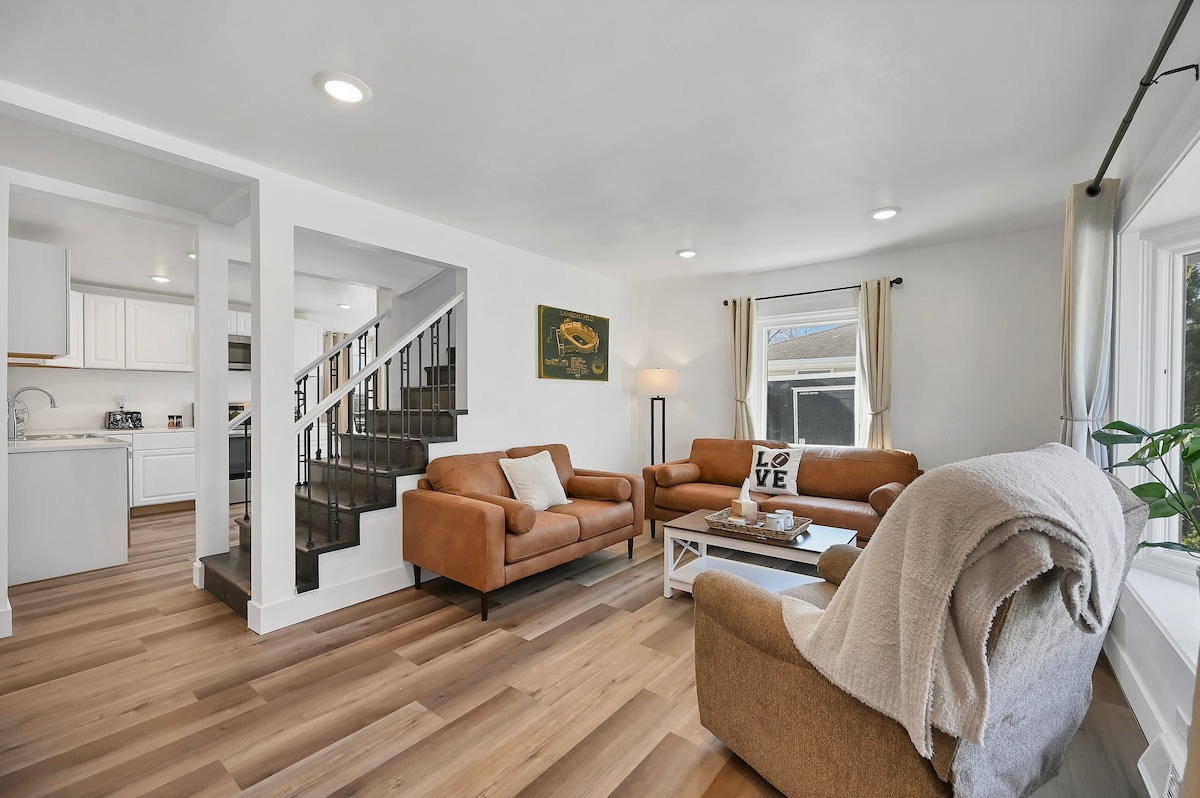 The living area features two brown sofas and a light-colored armchair situated around a coffee table. A brightly lit atmosphere is created by natural light entering through the windows. White walls and wooden flooring enhance the spaciousness of the room, with a staircase visible in the background.