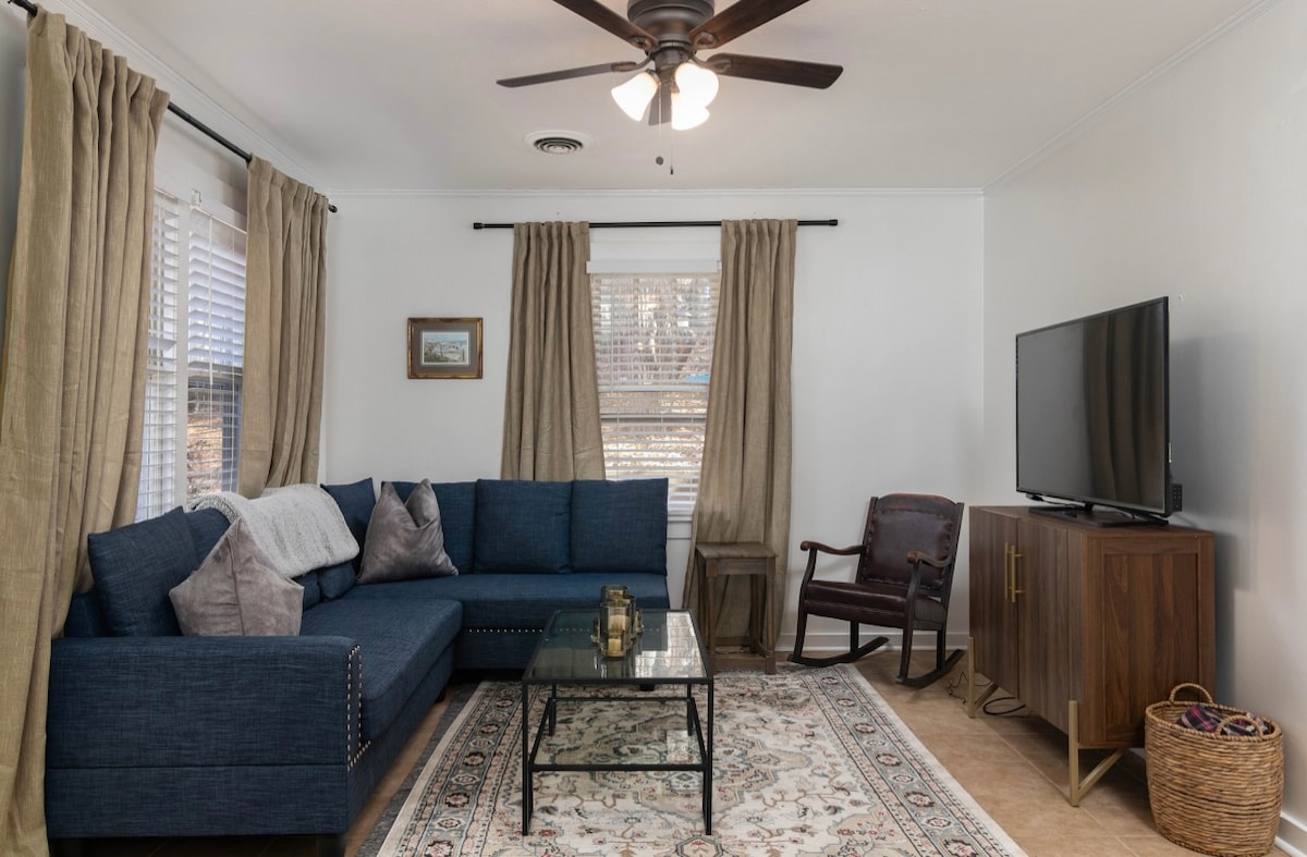 A living room is showcased, featuring a blue sectional sofa adorned with decorative pillows. A glass coffee table sits on a patterned area rug. Natural light filters through the window curtains, and a television is placed on a wooden cabinet. A comfortable armchair complements the space.