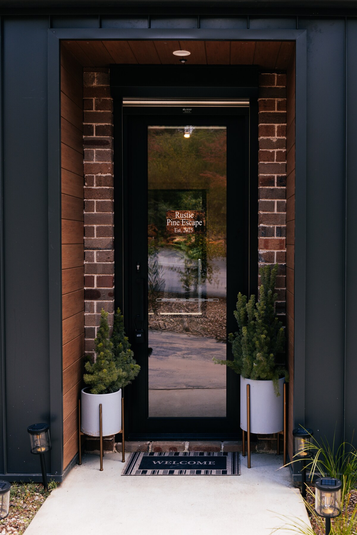 The entrance features a stylish door framed by modern brick and wood accents. On either side, potted plants add greenery, while a welcome mat is positioned on a smooth pathway leading to the entrance.