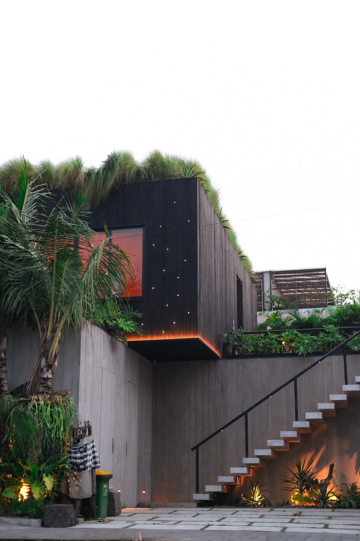 A modern structure features a green rooftop adorned with lush grass. Large windows offer glimpses of the interior space, while a staircase with slender railings leads to an entrance framed by diverse greenery. Soft lighting accentuates the building's design at dusk.