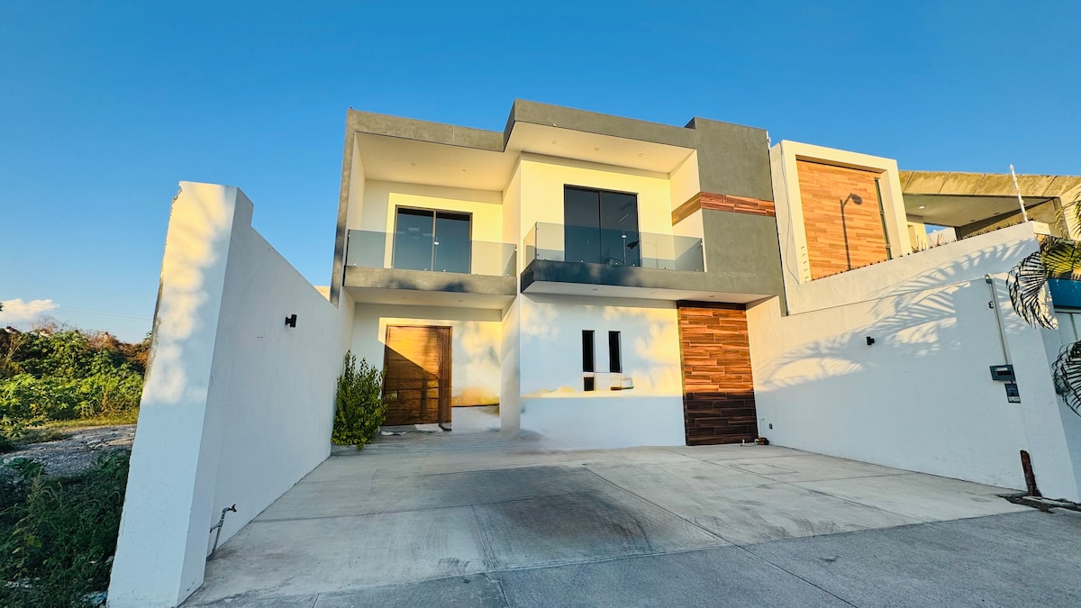 A modern two-story house is presented with a sleek facade, featuring large glass doors that offer a view of the interiors. A spacious driveway is visible in front, bordered by a low wall that complements the clean lines of the structure.
