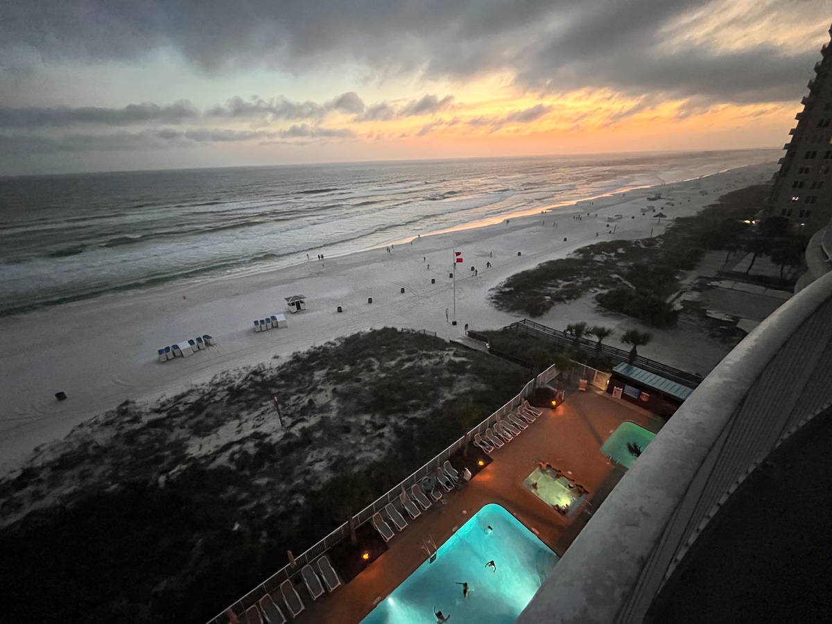 An expansive view of the beach at sunset is captured, revealing the gentle waves and sandy shore lined with umbrellas. The image features a heated swimming pool area below, illuminated by soft lights, with the ocean's horizon blending into the dusky sky.
