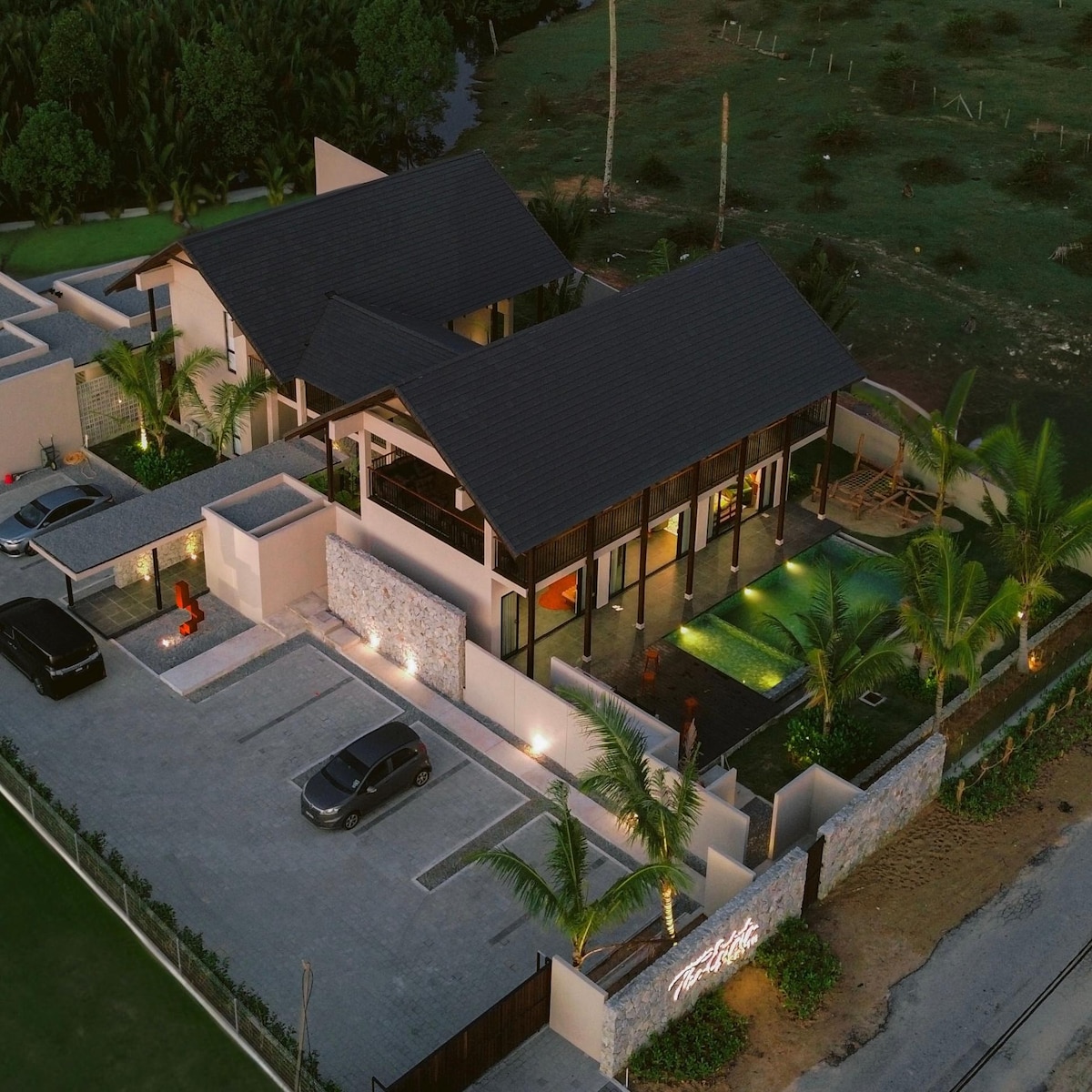 An aerial view of a modern estate showcases spacious outdoor areas with palm trees surrounding a tranquil pool. The multi-level building features a dark roof and expansive glass walls, creating a connection to the lush landscape and nearby greenery.
