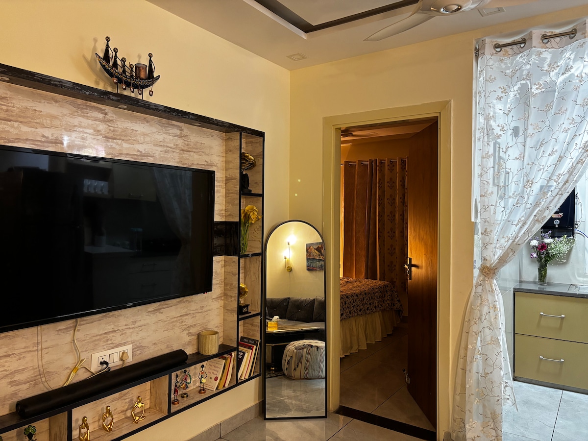 A living area displays a large wall-mounted television, accompanied by decorative shelves. An arched mirror stands beside the entryway, offering a view of a softly lit bedroom through an open door. Light-colored curtains drape around the kitchen space, adding a touch of warmth.
