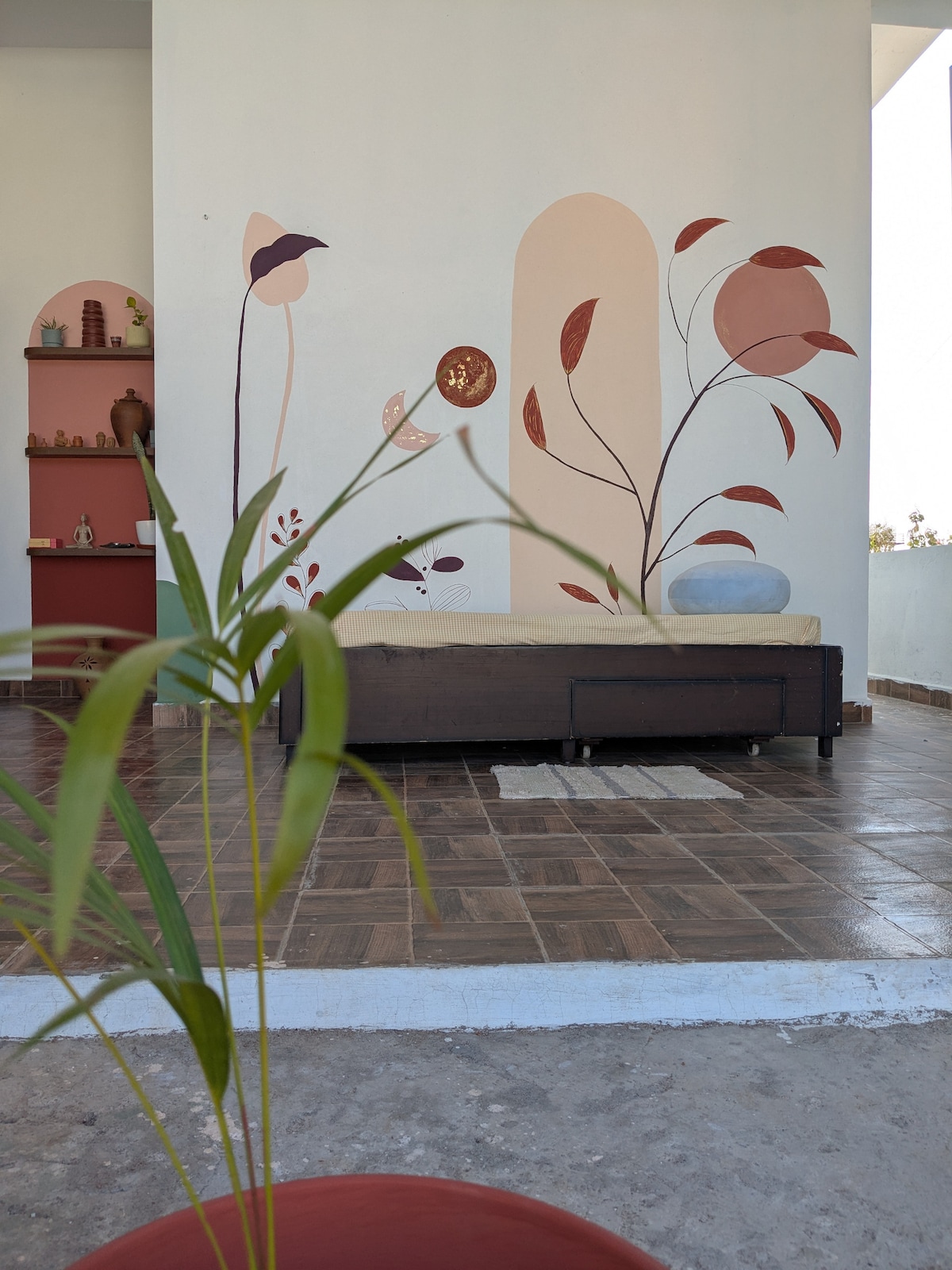 A cozy seating area features a simple couch against a wall adorned with abstract floral motifs in warm hues. Decorative shelves showcase various small art pieces. A potted plant adds a touch of greenery, with a tiled floor providing texture to the space.