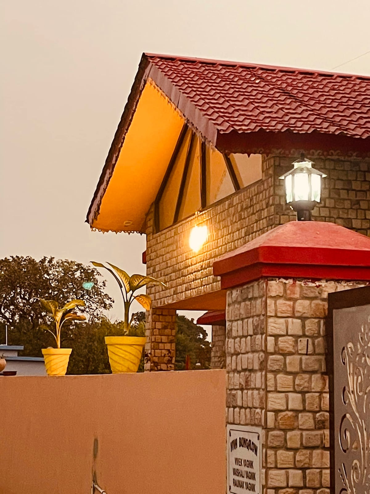 A charming villa exterior features a tiled roof with wooden accents and stone wall detailing. Two bright yellow planters with greenery sit on the ledge, while a soft outdoor light emits a gentle glow, enhancing the inviting entrance.