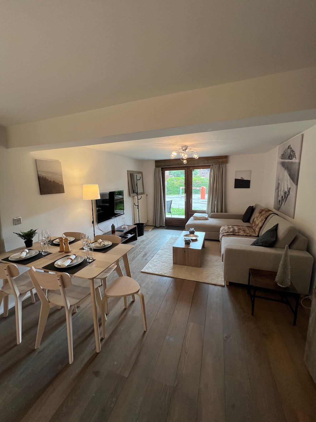 An open living area features a dining table set for four, complemented by a cozy seating arrangement. Natural light enters through large windows, with soft furnishings and neutral tones creating a serene space. The floor is wooden and enhances the room's warmth.