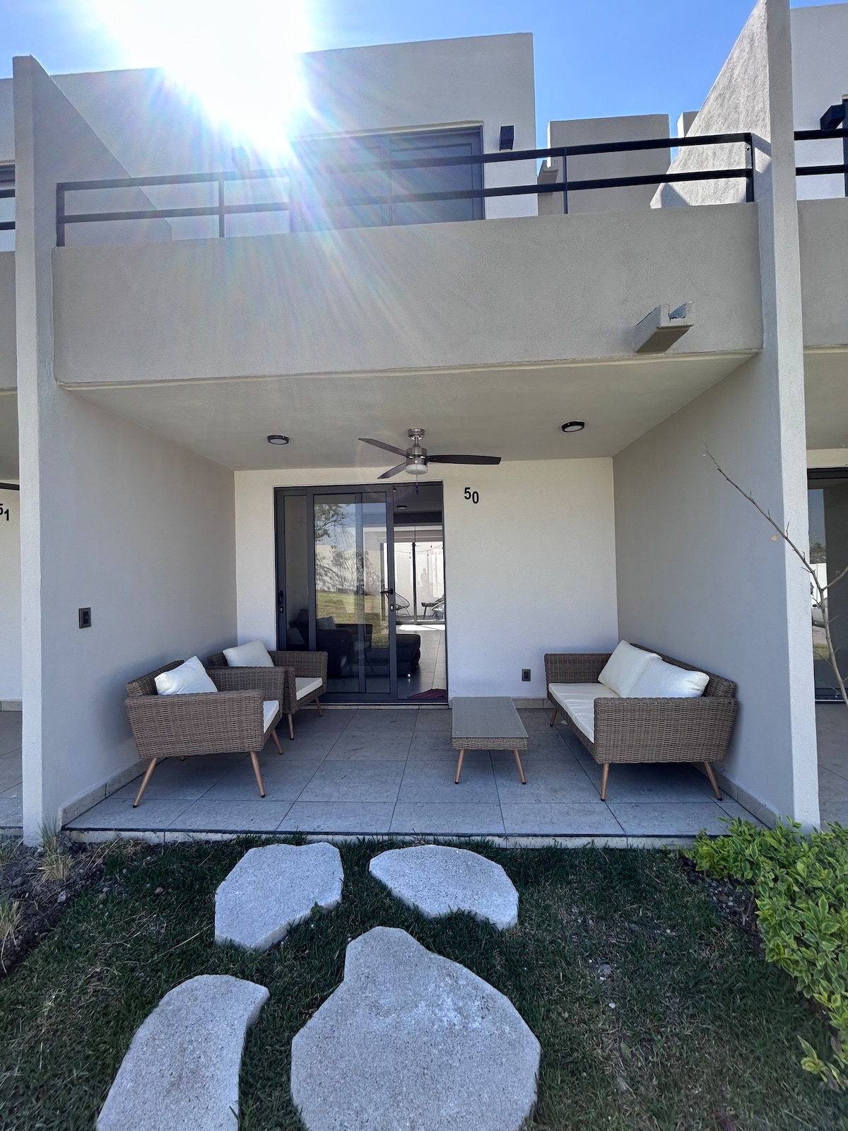 A private outdoor seating area is highlighted, featuring a table surrounded by four comfortable chairs. The space is framed by a light-colored wall and a ceiling fan is visible above. Sunlight filters in, illuminating the area, which is accessible via large glass doors.