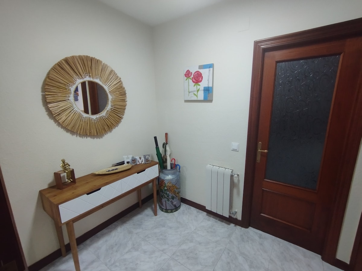 An entryway features a wooden console table with a decorative plate and a small trophy. A circular mirror with a woven frame hangs above, while a wall artwork depicting roses adds a touch of color. The door is made of wood with a frosted glass panel.