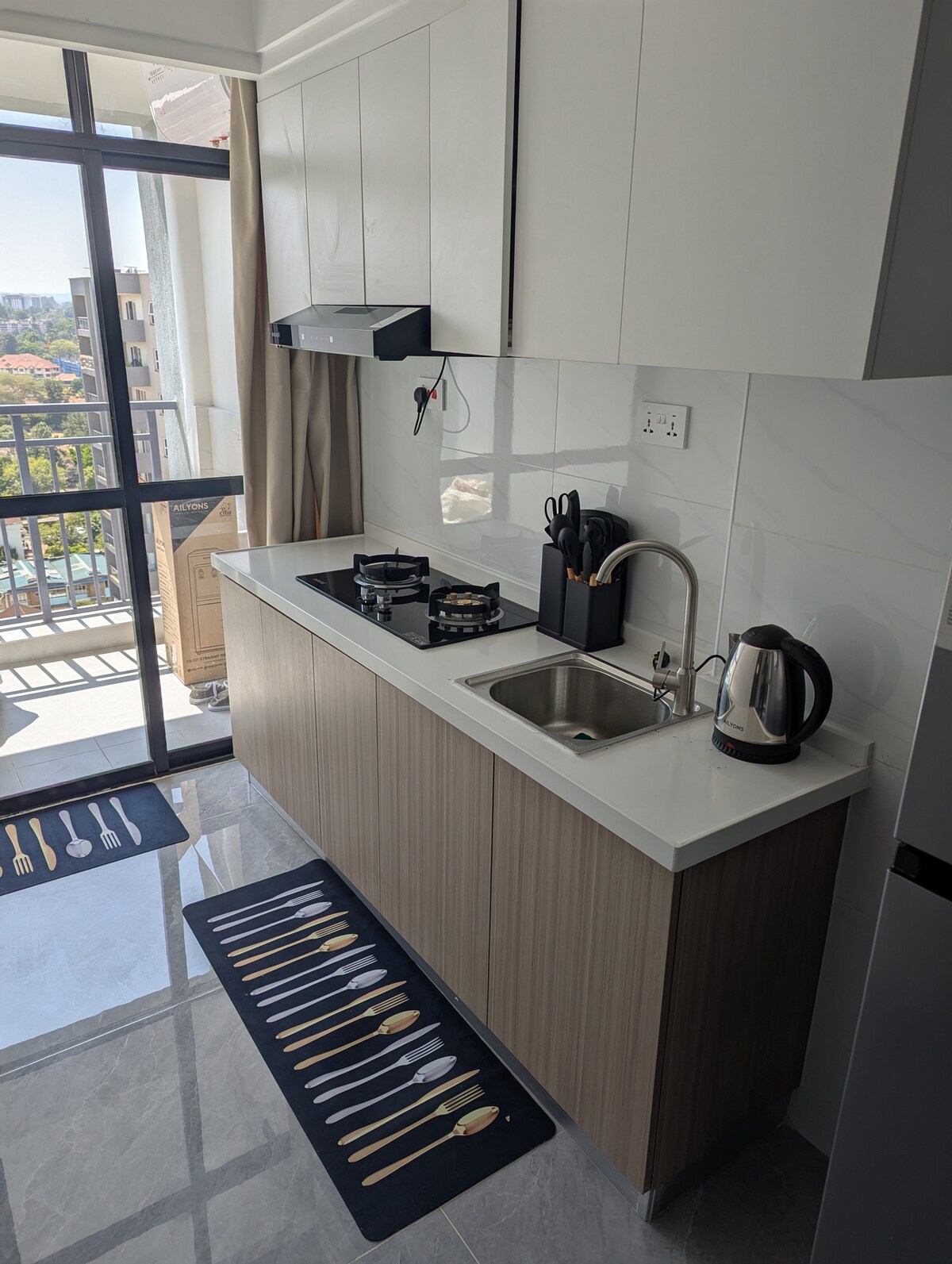 A modern kitchen area is equipped with sleek cabinetry and a white countertop. Two gas burners are positioned next to a stainless-steel sink. A kettle is placed beside utensils, with a decorative rug featuring utensil designs on the tiled floor. Large windows provide natural light.