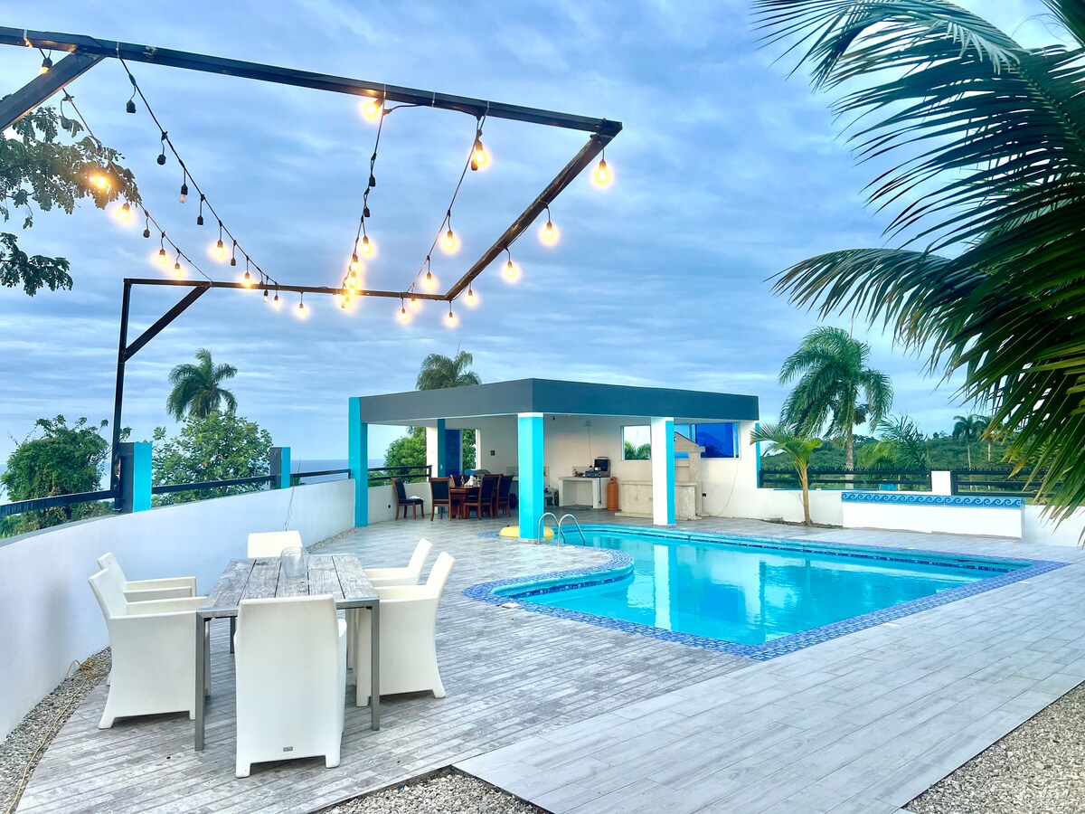 An outdoor patio features a spacious pool surrounded by a wooden deck. String lights hang overhead, illuminating the area. A dining table with six chairs is positioned nearby, and a shaded cooking area is visible in the background, nestled among lush greenery.