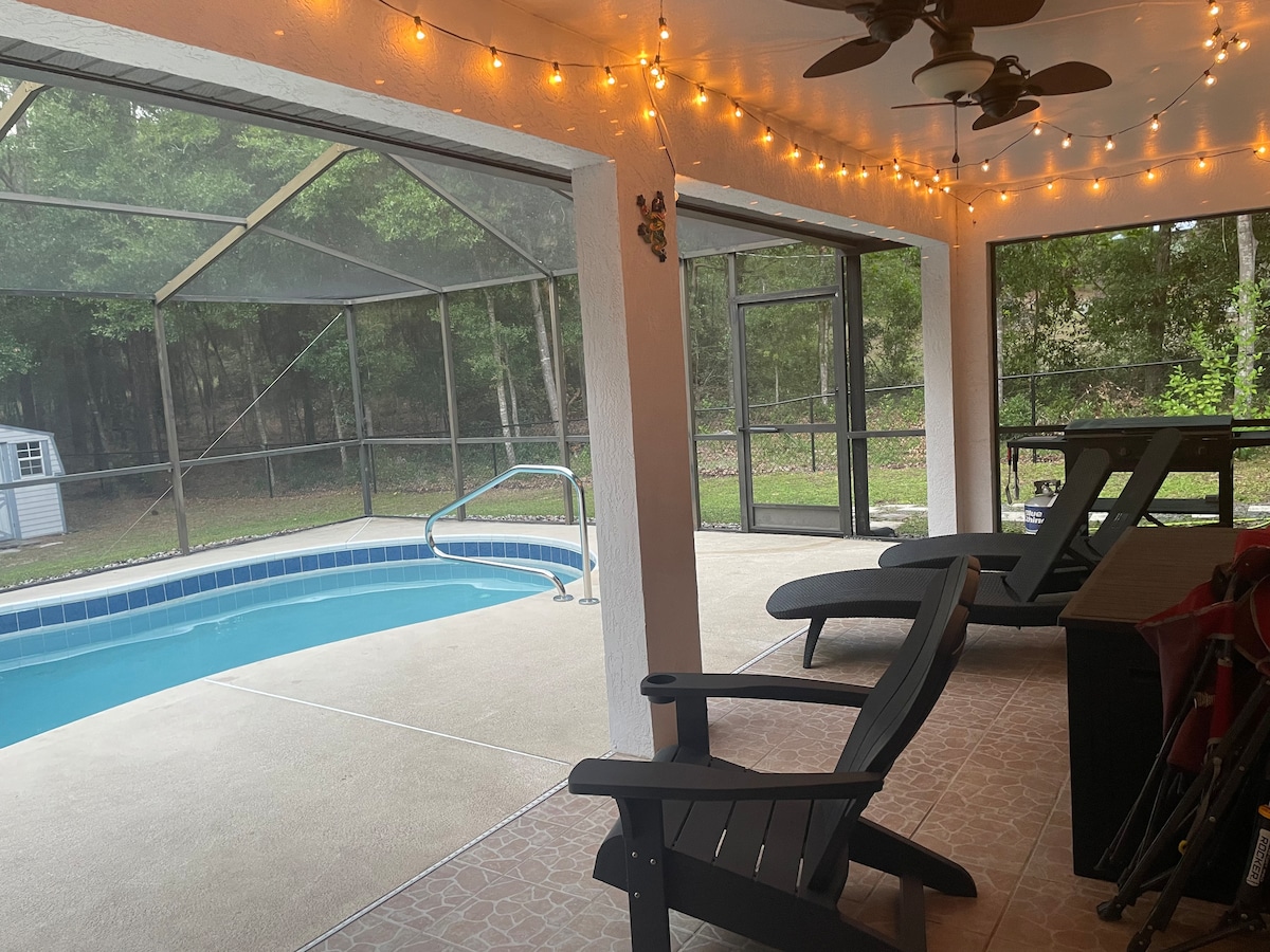 A screened-in lanai features a saltwater pool with a gently curving edge and a handrail. Lounge chairs are positioned beside the pool, with a shaded sitting area visible in the background. Warm string lights create a cozy ambience above the pool area, surrounded by lush greenery.