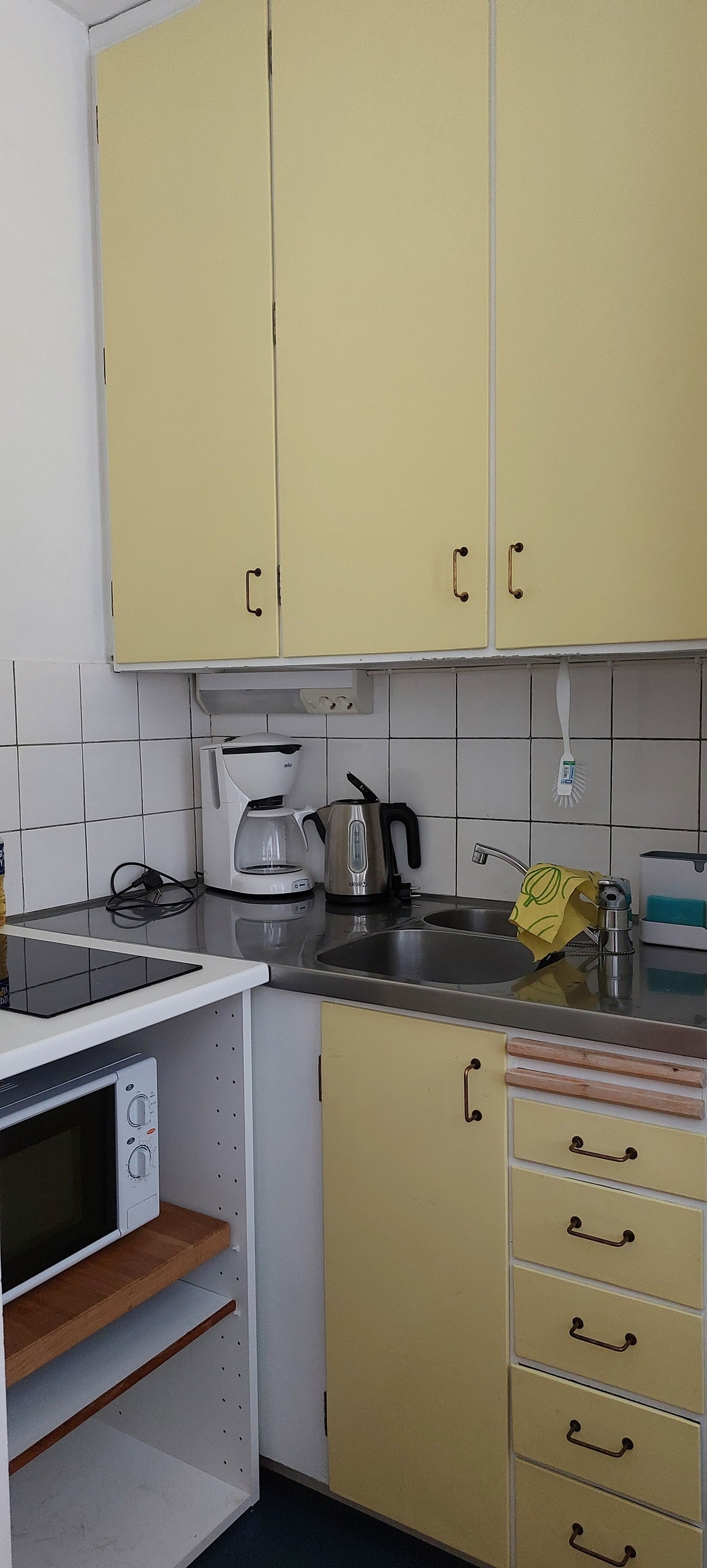The kitchen area features yellow cabinetry and a stainless steel countertop. A coffee maker and electric kettle are on display, along with a sink and a microwave. A small cutting board and dish towels are also present, suggesting a functional space for basic meal preparation.