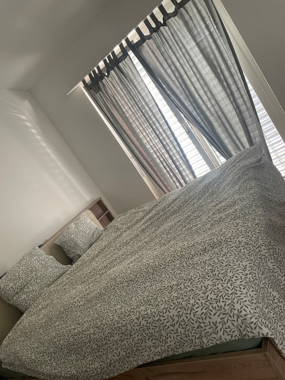 A spacious bed is covered with a patterned duvet, flanked by two textured pillows. Natural light pours in through light-filtering curtains, casting gentle shadows on the wall. The minimalist decor is complemented by a warm wooden bed frame.