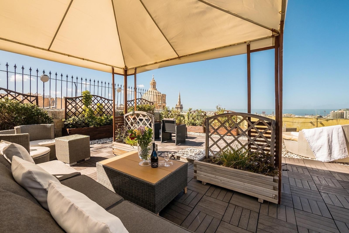 A spacious rooftop terrace features comfortable seating arrangements under a canopy. A wooden table is positioned at the center, accompanied by decorative plants and a bottle of wine. Scenic views of nearby historic buildings and the sea are visible in the background.