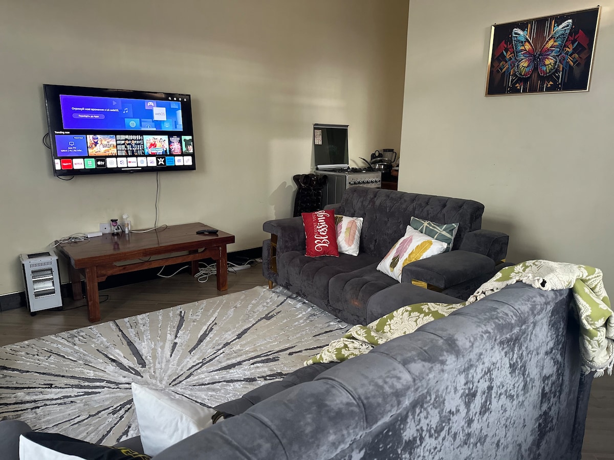 A comfortable living area is equipped with a large television mounted on the wall. An inviting grey sofa set surrounds a decorative area rug, while a wooden coffee table adds functionality. A piece of wall art featuring butterflies enhances the vibrant atmosphere.