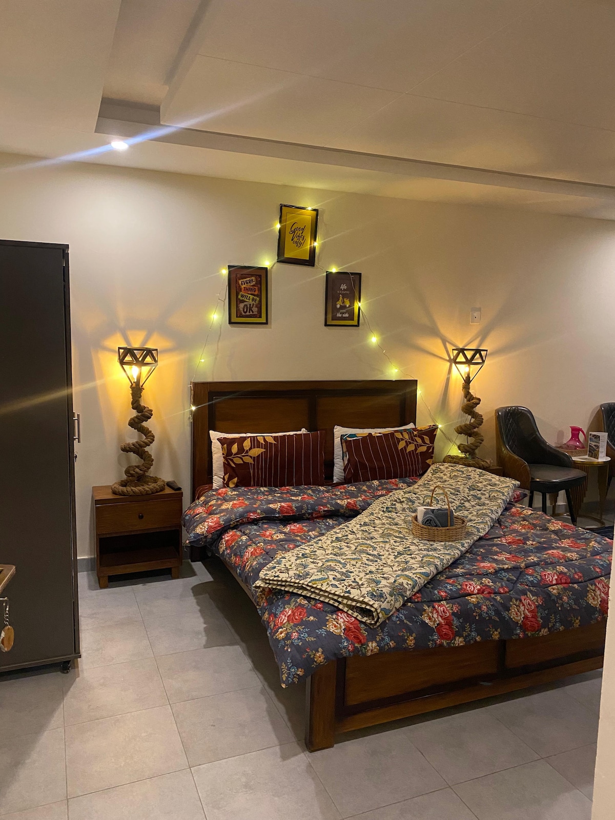A cozy sleeping area features a wooden bed adorned with floral bedding and decorative throw pillows. Two unique lamps provide soft lighting. A dark wardrobe is positioned nearby, and a small bedside table holds a decorative item. Light string accents add a warm glow to the space.