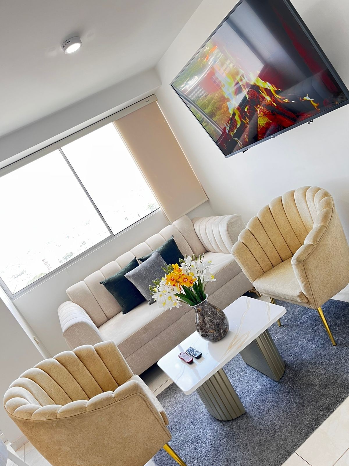 A modern living area features a plush beige sofa paired with two matching accent chairs. A small table sits centered, adorned with yellow flowers in a vase. A flat-screen TV is mounted on the wall, while a large window allows natural light to enter the space.