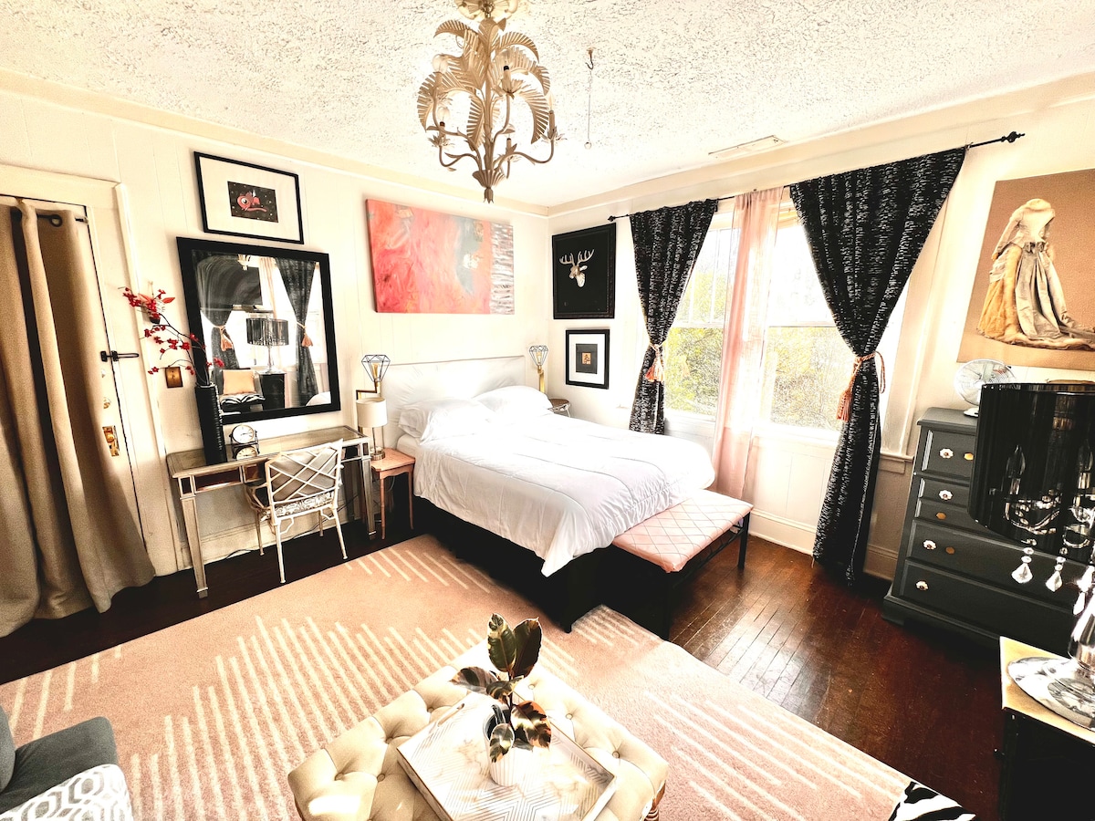 A bright bedroom features a comfortable bed covered with crisp white sheets. An elegant chandelier hangs from a textured ceiling. Large windows adorned with dark curtains allow natural light to filter in. Decor includes framed artwork and a stylish side table.