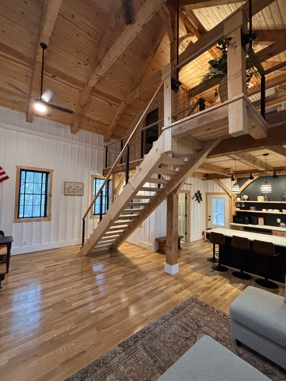 A spacious interior showcases a beautiful wooden staircase leading to an open loft. Natural light streams through the windows, illuminating the hardwood floors. The cozy living area is complemented by a neutral-toned couch and a decorative area rug, enhancing the room's inviting atmosphere.